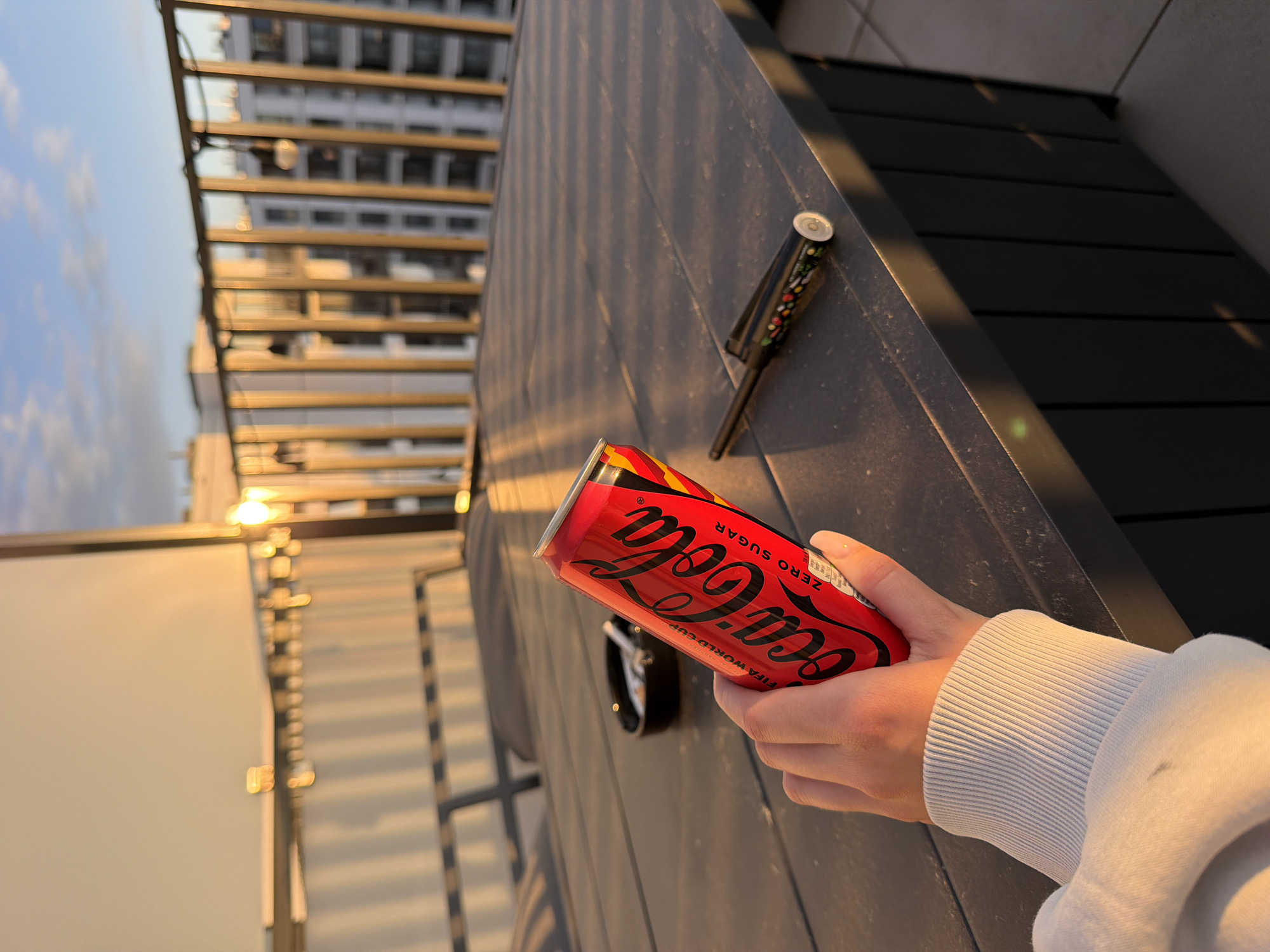 A hand in a white sweater holds a red can of Coca-Cola Zero Sugar on a black surface. A black object is next to the can. In the background, there is a building. The image appears to be taken on a rooftop. The location is a city, but it is not specified.