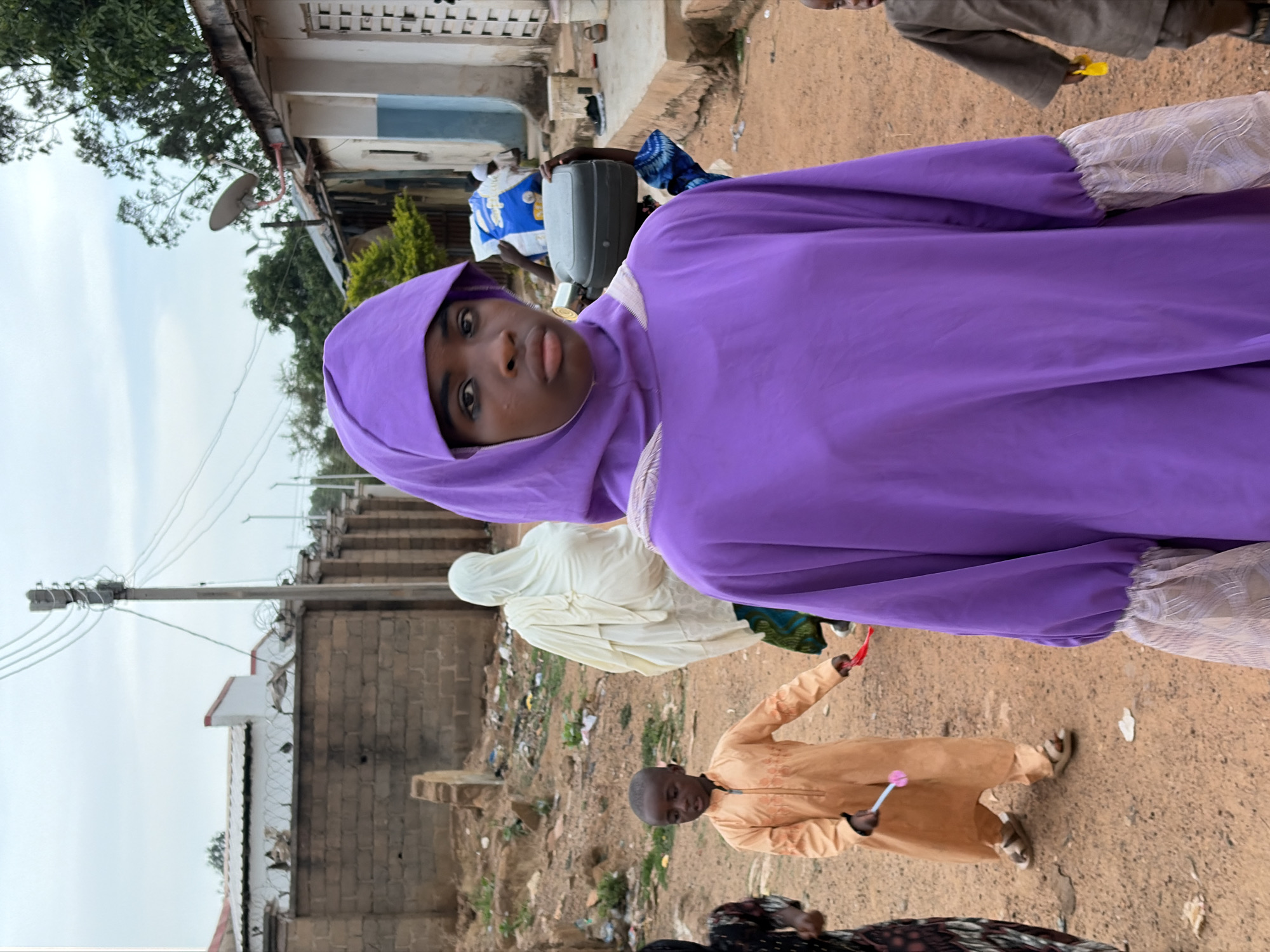 The image, taken in Barakin Lerawi, Nigeria, shows a young woman in a purple hijab and dress as the primary subject. Other individuals are visible in the background: a person in a white hijab and a child in tan clothing holding a small toy. The setting appears to be a street or open area with scattered trash and vegetation. A building is visible in the background along with power lines.