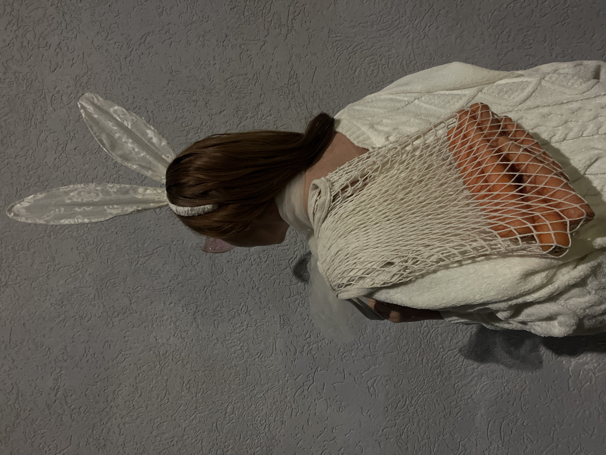A person with brown hair wearing white lace bunny ears stands in front of a gray textured wall. They are wearing a white towel and holding a white net bag with a hand visible inside. The location is not discernible from the image.