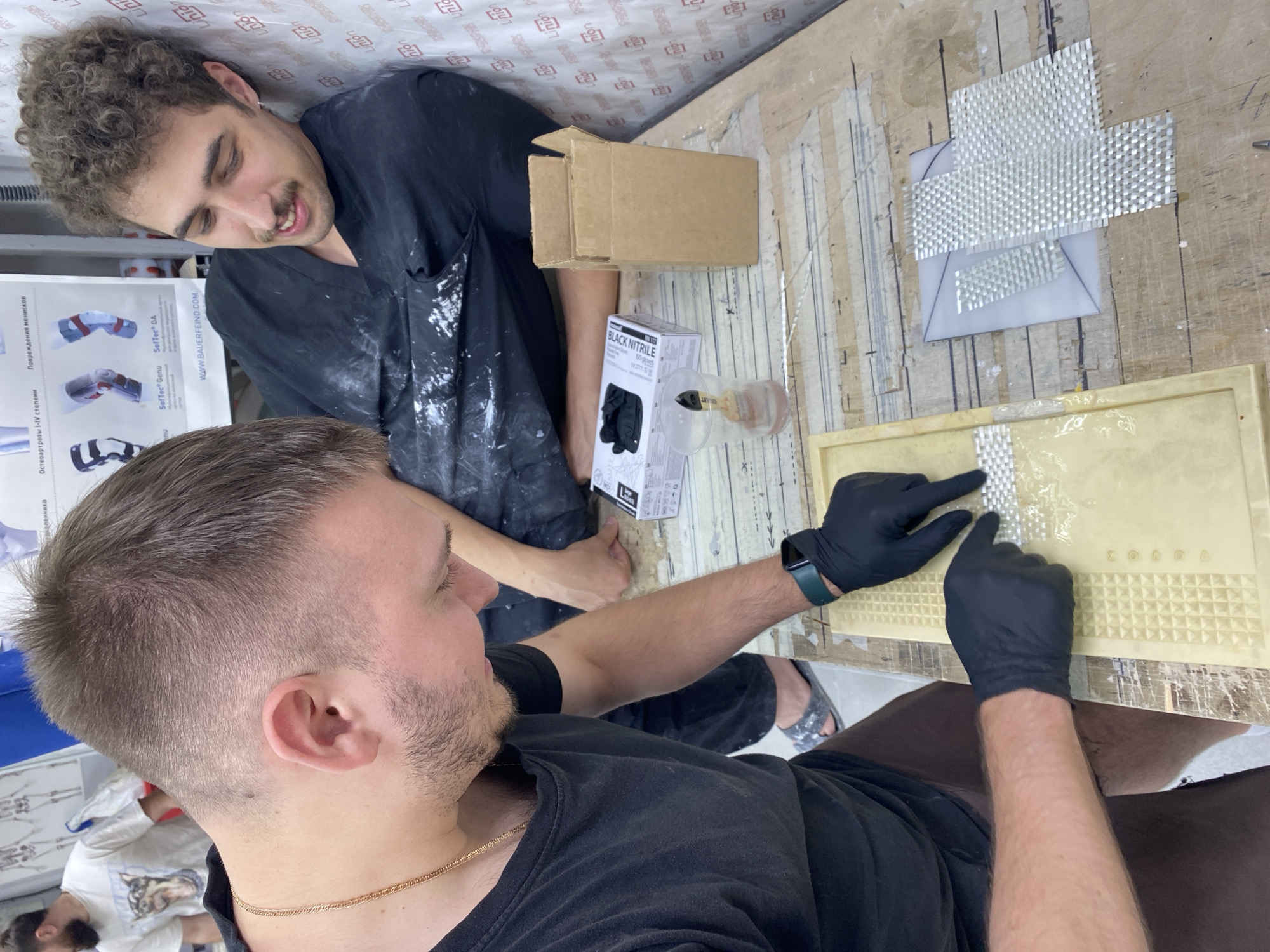 The image depicts two individuals engaged in what appears to be a craft or repair activity. One person, in the foreground and to the left, wears a black t-shirt and black gloves. They are actively working on a light-colored, textured surface, using their gloved hands to manipulate small, white, grid-like pieces and a clear liquid. A gold chain is visible around their neck. The other person is partially visible in the background, leaning against a wall covered with patterned material. This person also wears dark clothing and has curly hair and a mustache.
On the table, in addition to the work surface, are various objects: a clear plastic cup with a brush and liquid, a cardboard box, a package labeled "BLACK NITRILE," and several pieces of transparent, grid-patterned material. The background shows a wall with anatomical diagrams and text, suggesting a workshop or educational setting. The overall environment appears to be indoors, with a wooden table surface and a textured wall. There is no specific information in the image to indicate the location within the city of Vladimir, Russia.