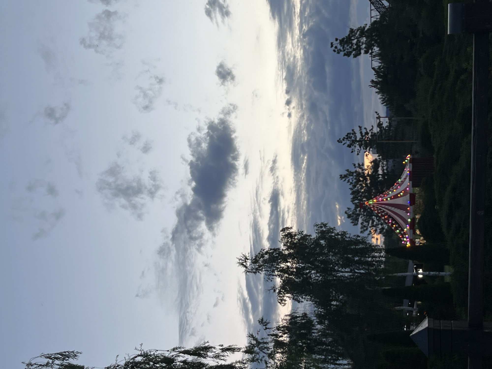 The image depicts a circus tent lit by colorful string lights, set against a twilight sky with scattered clouds. The tent is red and white striped. Surrounding the tent is dense green foliage and several trees. In the foreground, a dark wooden fence is visible, with a glimpse of a dark fence post and a small structure resembling a mailbox. The overall atmosphere is calm and serene, suggesting a rural or park-like setting. The scene does not contain any people or discernible actions. The specific location in the city of Dnipro, Ukraine, cannot be determined from the visual information provided in the image.