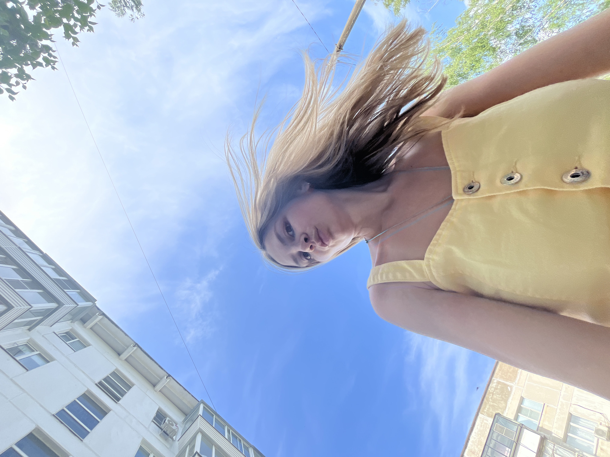 A low-angle shot of a young woman with fair skin and blonde hair, wearing a yellow top with buttons. Her hair is partially obscuring her face.
Behind her is a blue sky with scattered white clouds, and segments of buildings, trees, and a wire.
This photograph was taken outdoors in the city of Chaikovsky, Russia.
