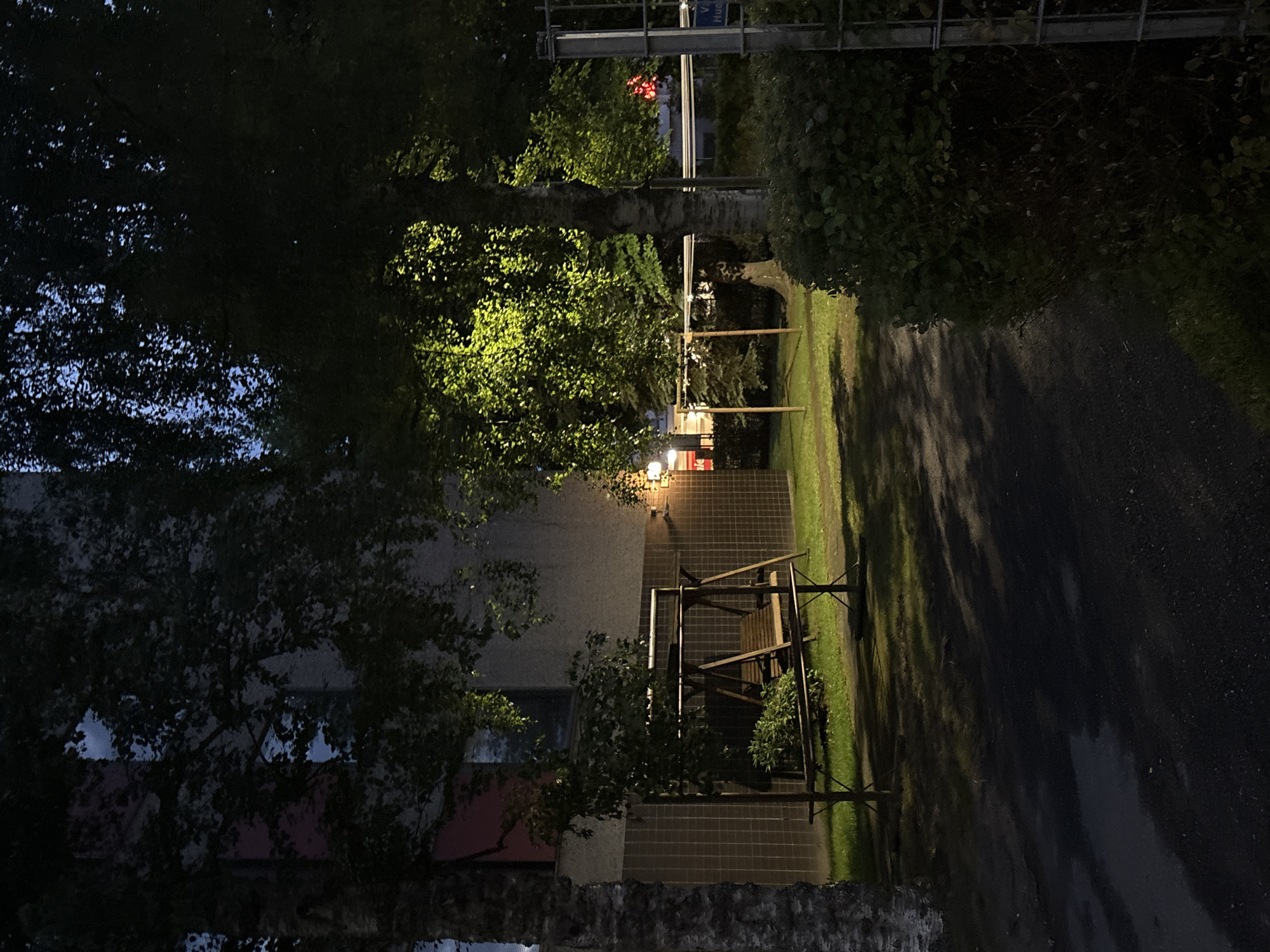 The image depicts a courtyard area in the city of Pori, Finland, at dusk or during the night. In the foreground, a paved walkway is visible, leading towards a grassy area where a wooden swing set with a bench is situated. Behind the swing set, a building with a light-colored, tiled facade is visible. Trees with lush green foliage are scattered throughout the scene, with one large tree in the mid-ground illuminated by a light source, casting a bright yellow glow on its leaves. A dense hedge is located on the right side of the image, partially obscuring a metal fence. The overall atmosphere is dark and somewhat muted, with artificial lighting highlighting specific areas.