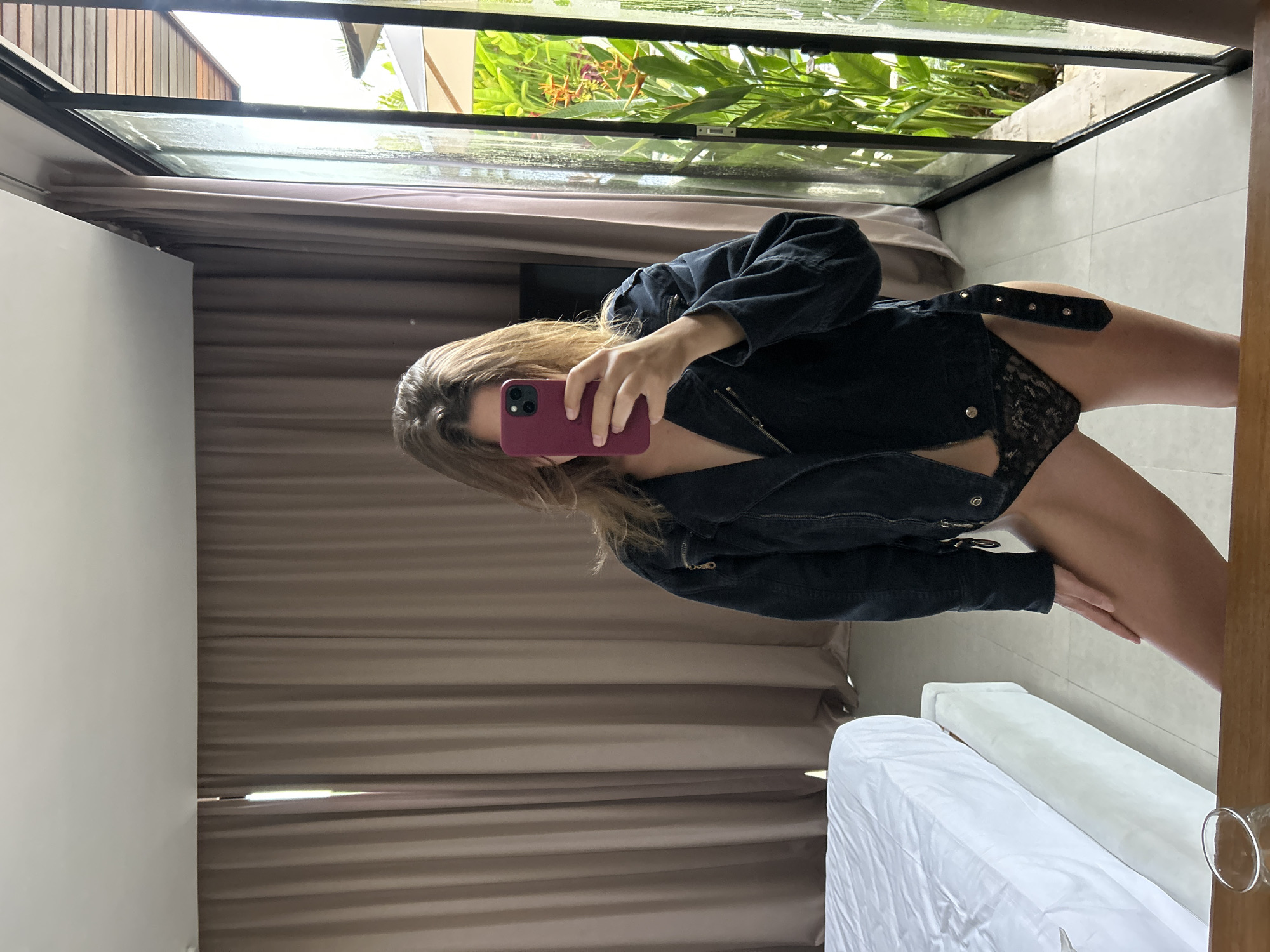 A woman with long brown hair is holding a red cell phone up to her face and taking a selfie. She is wearing a black denim jacket and black lace underwear with a studded belt. The setting appears to be a room in a home or hotel, with a large window showing lush greenery, possibly located in the Ubud District of Indonesia, based on the vegetation and architectural style. Behind the woman are long, brown curtains. A white bed is partially visible in the foreground. The room appears to be well-lit.
