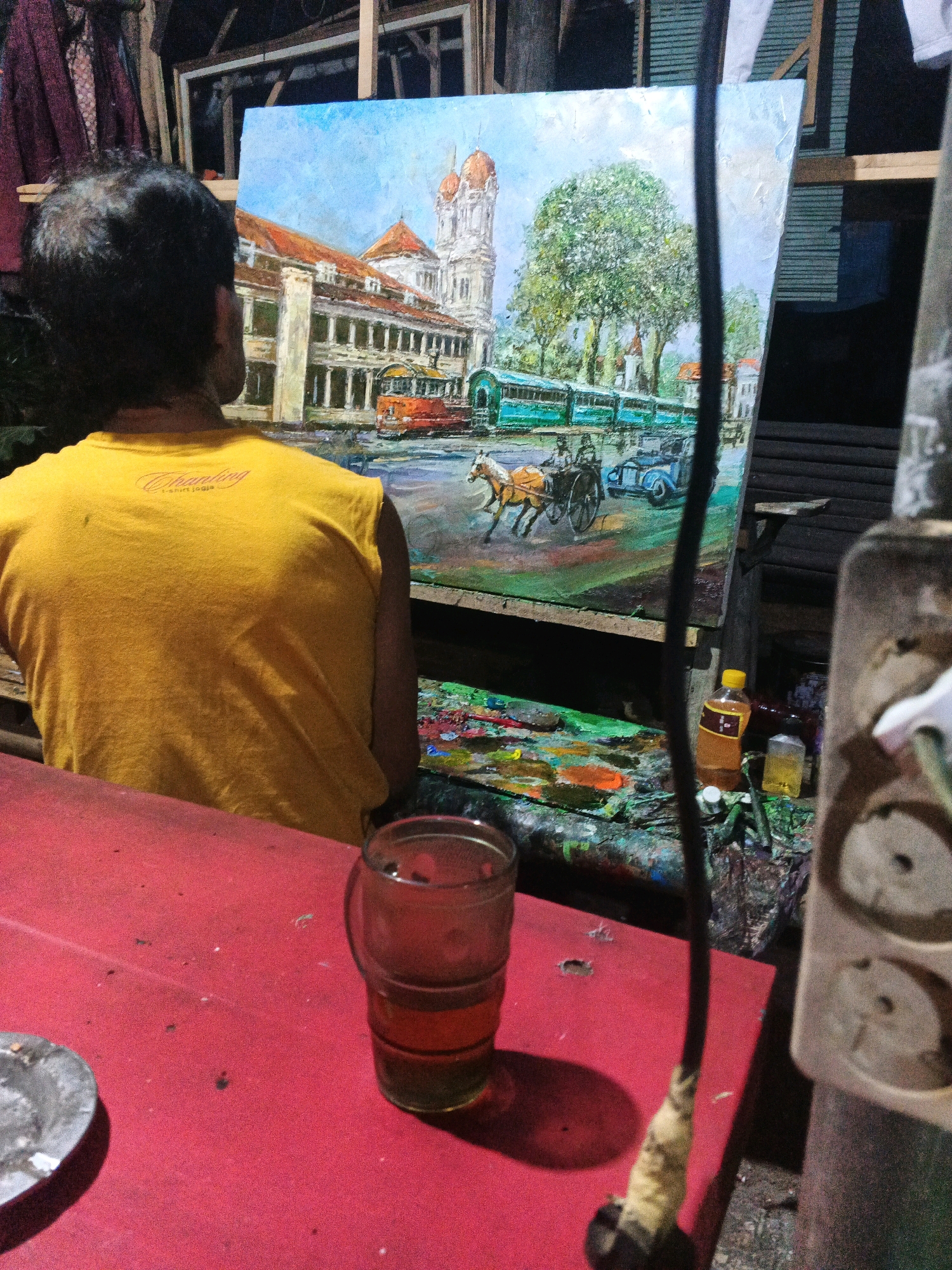 The image depicts a scene in Ungaran, Indonesia, where an artist is working on a painting. The artist, seen from behind, is wearing a yellow t-shirt and is focused on a large canvas on an easel. The painting itself is a landscape featuring colonial-era architecture, a tram, a horse-drawn carriage, and trees under a bright blue sky.
In the foreground, a red table holds an ashtray and a glass of amber-colored liquid. To the right, part of an electrical outlet is visible. The background reveals a rustic setting, possibly an outdoor studio, with wooden structures and hanging fabrics. The overall atmosphere suggests a moment of artistic creation in a local Indonesian setting.