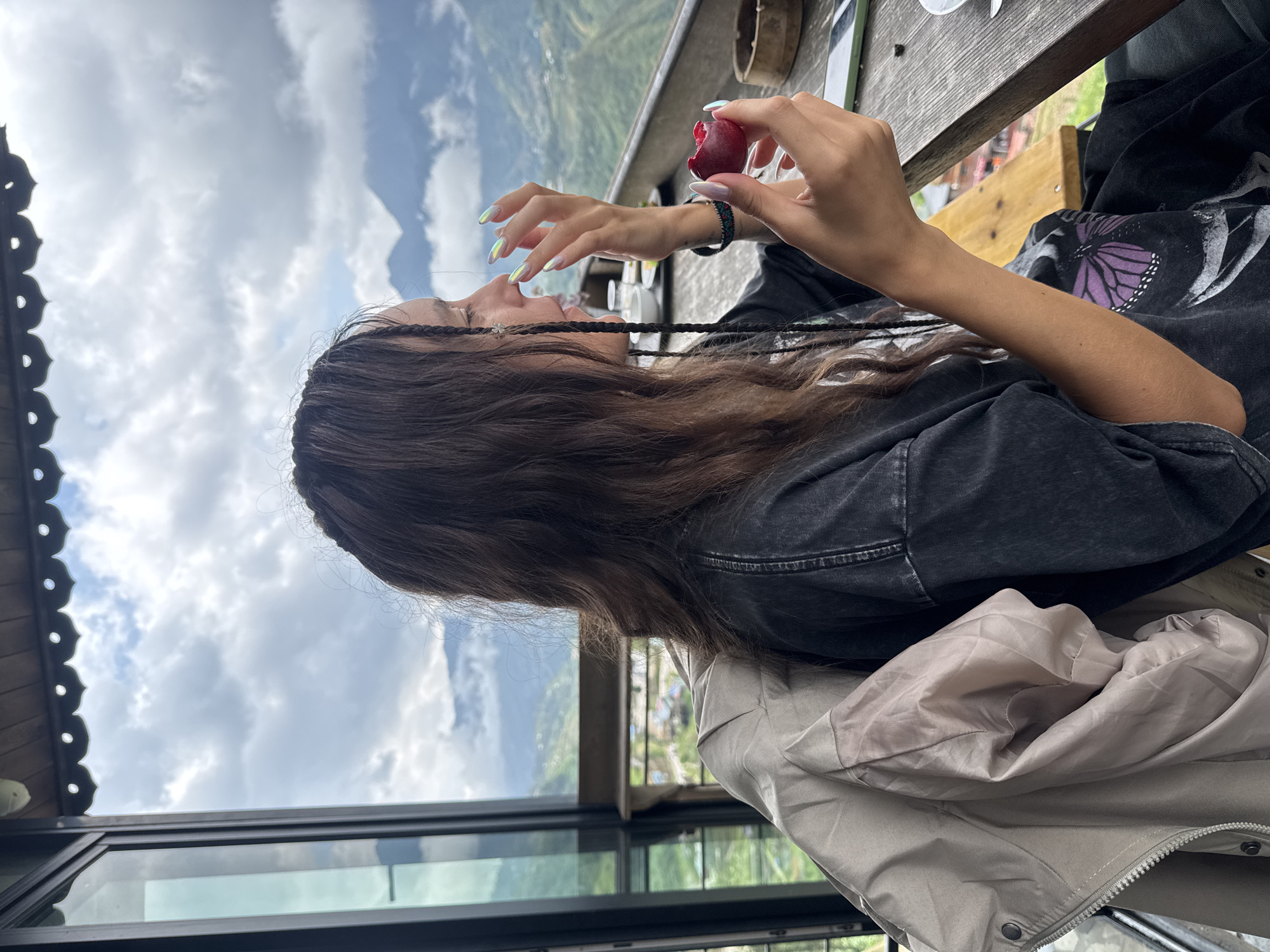 A young woman sits at a wooden table overlooking a mountain valley in Sa Pa Ward, Vietnam. She holds a red fruit in her right hand and is about to eat it. She wears a black T-shirt with a purple butterfly graphic and a light gray jacket. Her hair is long and brown, with two small braids. The table has a few dishes and utensils on it. The background shows a mountainous landscape with a cloudy sky. A building with large windows can be seen below the table.