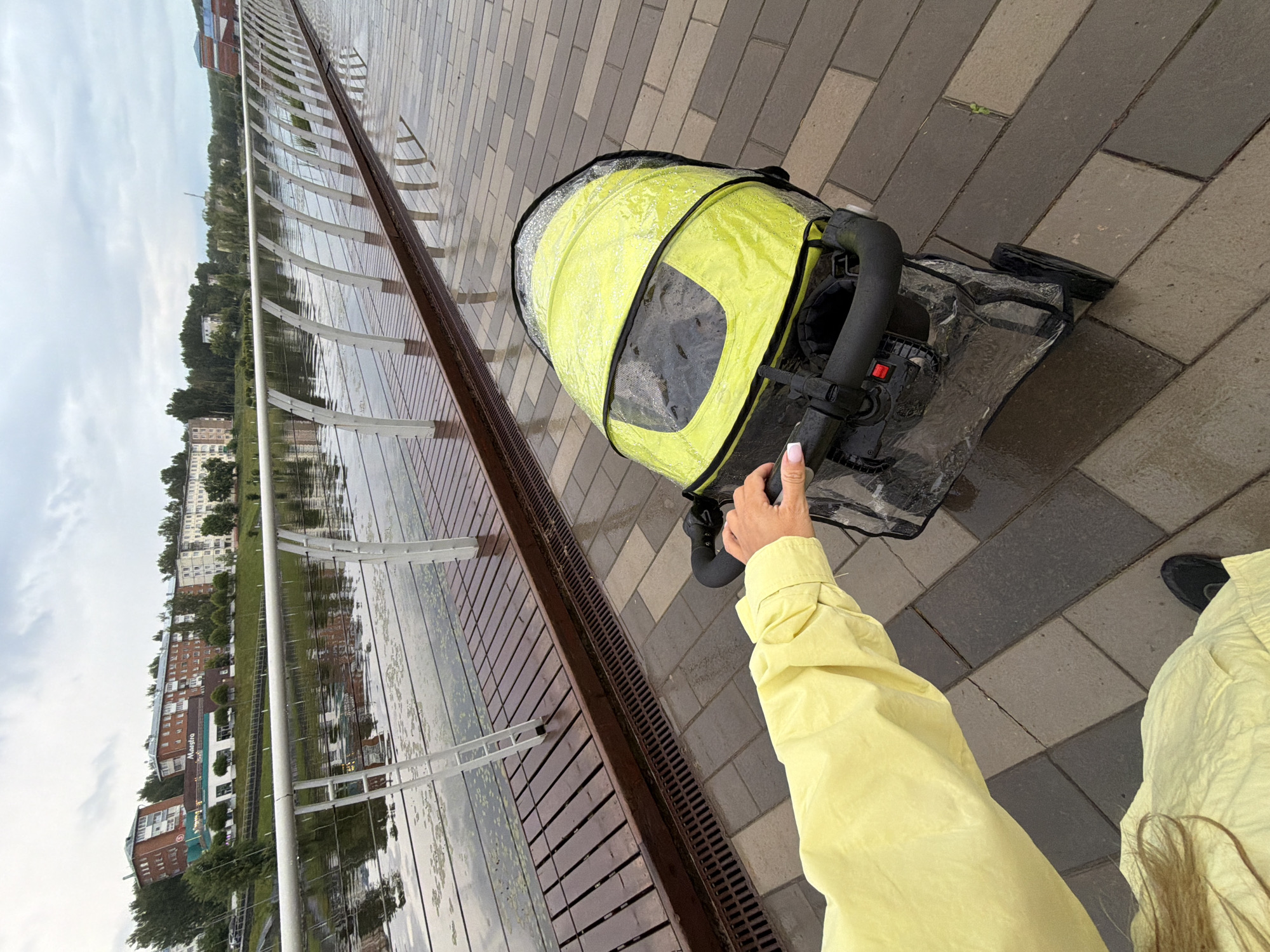 A person is seen from a first-person perspective, pushing a stroller along a wet, paved promenade in Votkinsk, Russia. The individual's left arm, clad in a bright yellow long-sleeved jacket, extends into the frame from the lower left, gripping the black handle of the stroller. The person's left hand has light-colored nail polish. A section of long, light brown hair is visible over the left shoulder, and the tip of a black shoe is visible at the very bottom left.
The stroller has a vivid yellow canopy covered by a transparent plastic rain cover, on which water droplets are visible, indicating recent rain. A clear plastic cover also encases the lower section of the stroller. The stroller has black wheels and a black handlebar with a red button.
The ground beneath is a wet, tiled walkway composed of rectangular pavers in various shades of gray and brown, laid in an offset pattern. To the left of the walkway is a dark brown wooden boardwalk, also visibly wet, running alongside a body of water. A modern white railing with arched posts and horizontal cables separates the boardwalk from the water.
The body of water, likely a river or canal, appears calm, reflecting the overcast sky and distant structures. Patches of green aquatic vegetation float on its surface.
On the opposite bank, under an overcast sky, a cityscape of Votkinsk is visible. It features several multi-story residential buildings, primarily with red brick and lighter facades, interspersed with numerous trees. One prominent building on the far bank has a blue sign that reads "Maestro" on a white background, identifiable as part of the Maestro complex in Votkinsk. The wet surfaces and cloudy sky indicate damp, overcast weather conditions.