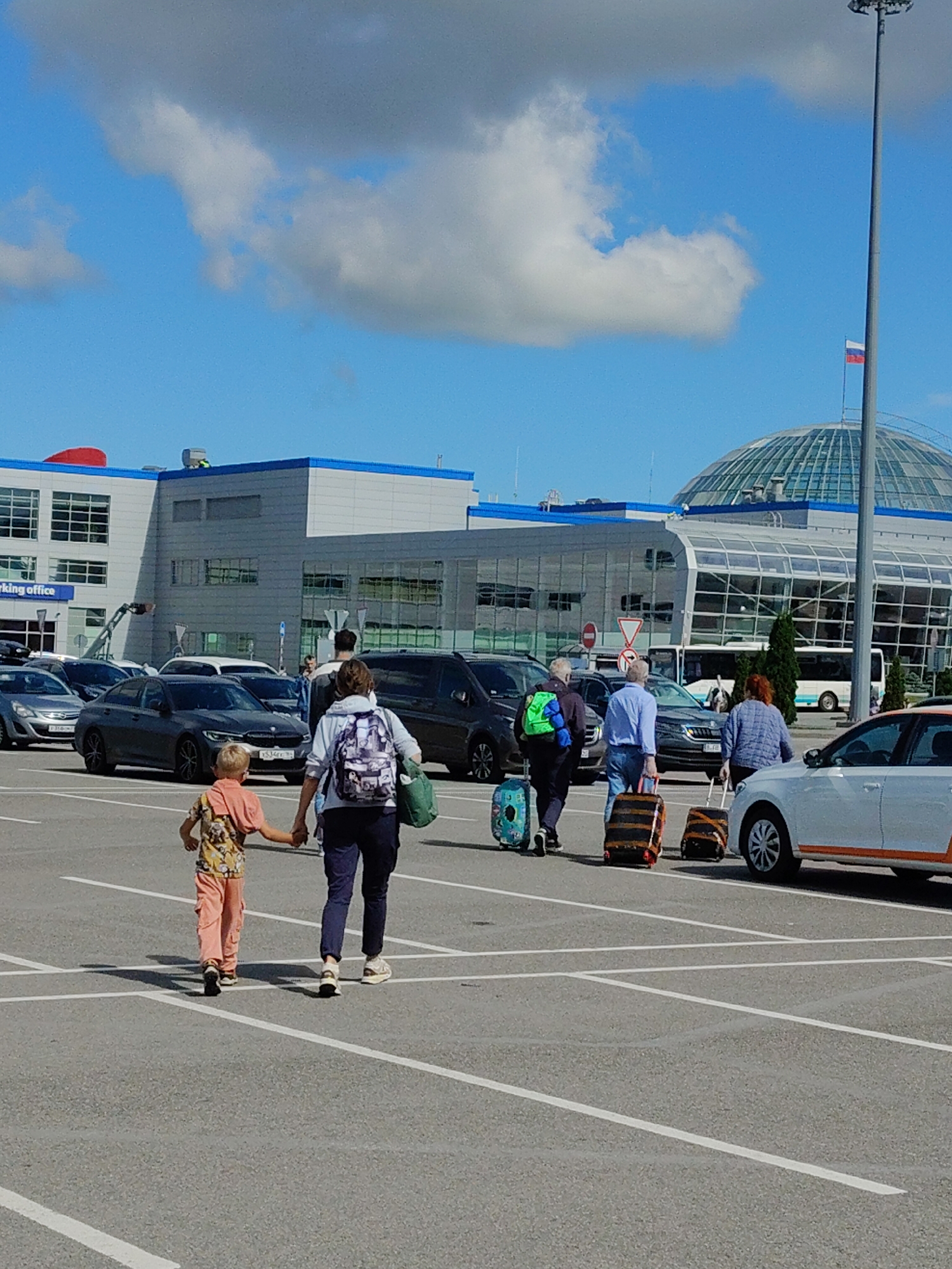 A group of people are walking across a paved parking lot towards a modern airport terminal. The terminal building has large glass windows and a distinctive dome-shaped roof. The sky is blue with scattered white clouds. Several cars are parked in the lot, and a white car with an orange stripe is on the right side of the frame. In the foreground, a woman is holding the hand of a young boy. The boy is wearing an orange patterned shirt and peach-colored pants. The woman is wearing a backpack. Behind them, other individuals are pulling luggage. The scene appears to be at Khrabrovo Airport in Kaliningrad Oblast, Russia, given the architecture and the Russian flag visible on a flagpole near the terminal.