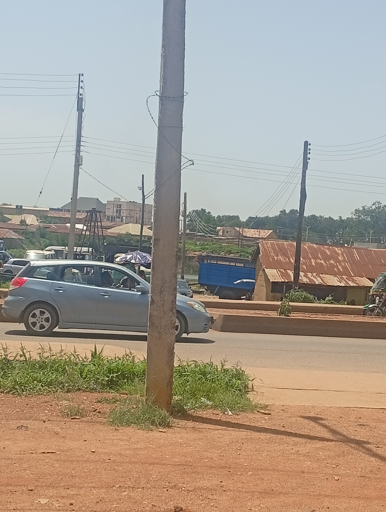 The image depicts a roadside scene in Bukuru, Nigeria. A light blue station wagon is parked on the left side of the road. In the background, there are several buildings with corrugated iron roofs, some appearing to be market stalls or residential structures. A blue truck is visible to the right of the station wagon. Power lines and utility poles run across the upper portion of the image, indicating an urban or semi-urban setting. Vegetation, including grass and some trees, is present along the roadside and in the background. The ground in the foreground is a reddish-brown earth. The sky is clear and bright, suggesting a sunny day.