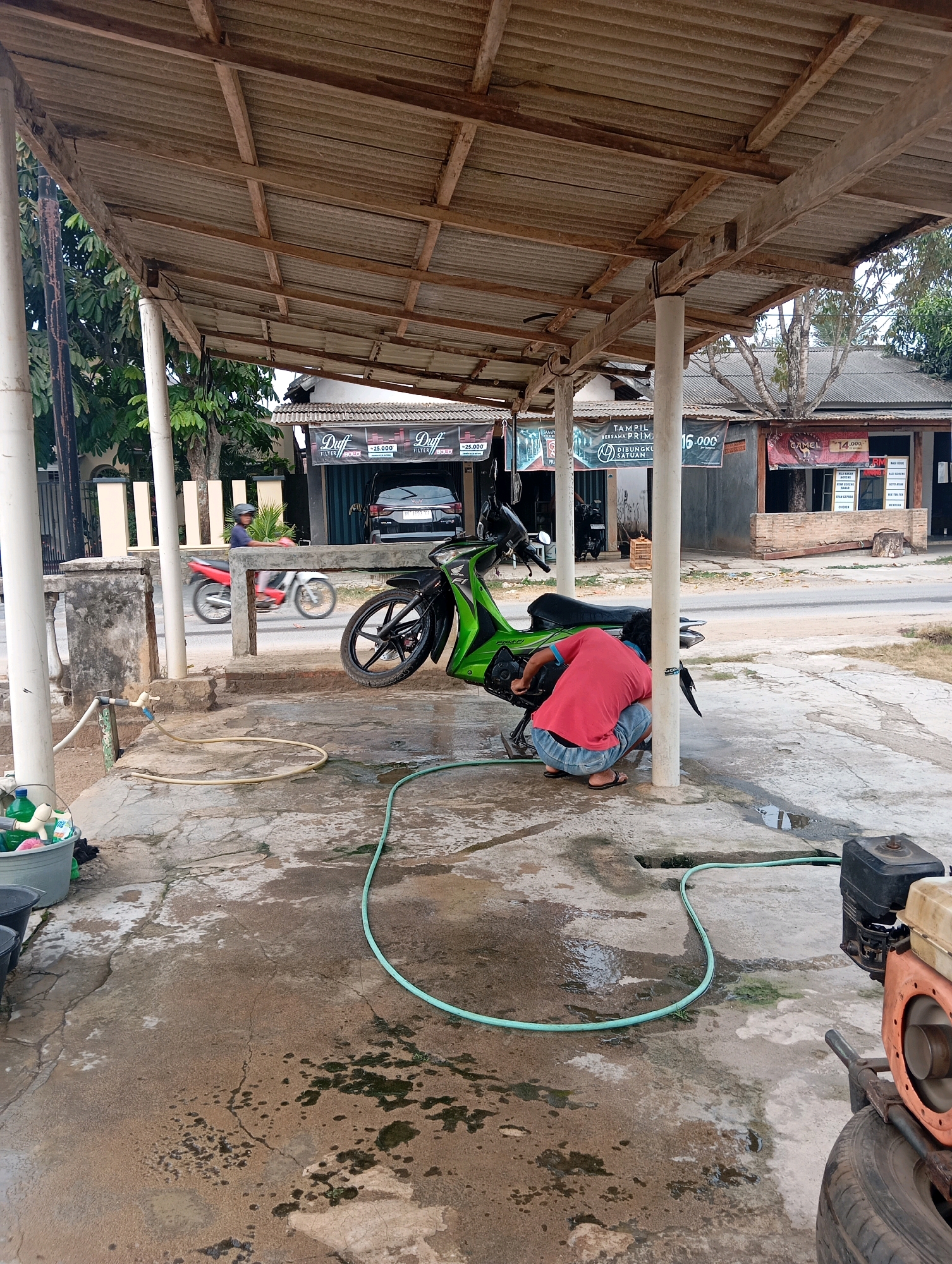 The image depicts an outdoor scene under a makeshift roof structure, likely in Leman Benawi, Indonesia. A person wearing a red shirt and blue shorts is crouching and working on a green and black motorcycle that is parked on a wet concrete surface. A green hose snakes across the ground, connected to a water source. To the left, a gray bucket and other cleaning supplies are visible. In the background, there are several storefronts with various signs, including one with "Duff" and "£25.000". A black car is parked in front of one of the buildings. A person on a red and white motorcycle is seen riding on the road in the middle ground. The environment appears to be a commercial or roadside area with buildings and a paved road.
