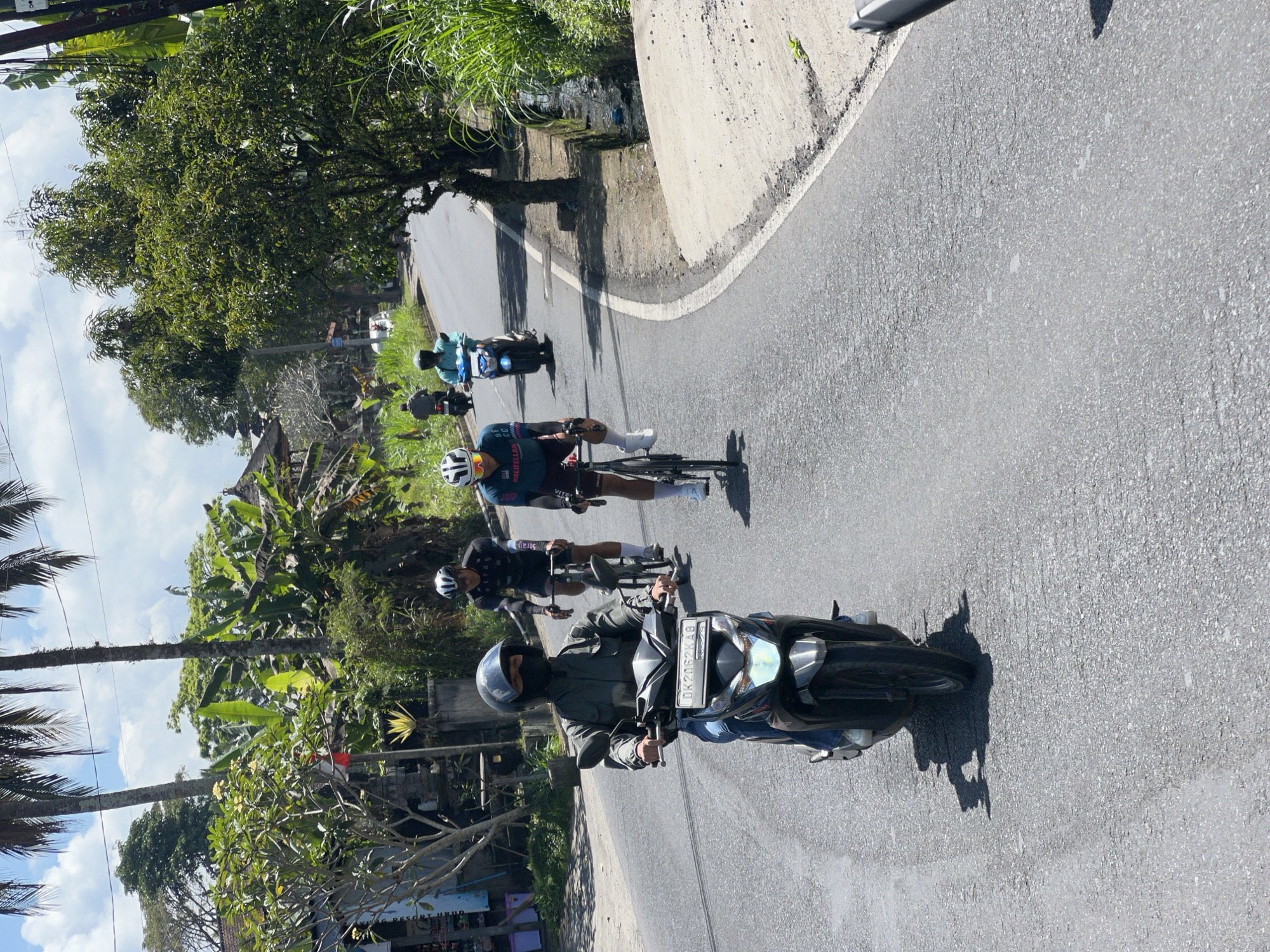 In Payangan, Indonesia, on a sunny day, a person rides a black scooter in the foreground of a paved road. The scooter has a license plate that reads "DK 2062 KAB". Behind the scooter, two cyclists are visible. The cyclist closest to the scooter wears a black helmet and a turquoise cycling jersey with white accents. The cyclist behind them wears a black helmet and a black long-sleeved cycling jersey with white lettering. Further in the distance, two more individuals are seen on motorbikes. The road is bordered by lush green foliage, including palm trees and banana trees, and a small ditch with water on the right side. Buildings and more trees are visible in the background. The sky is partly cloudy.
