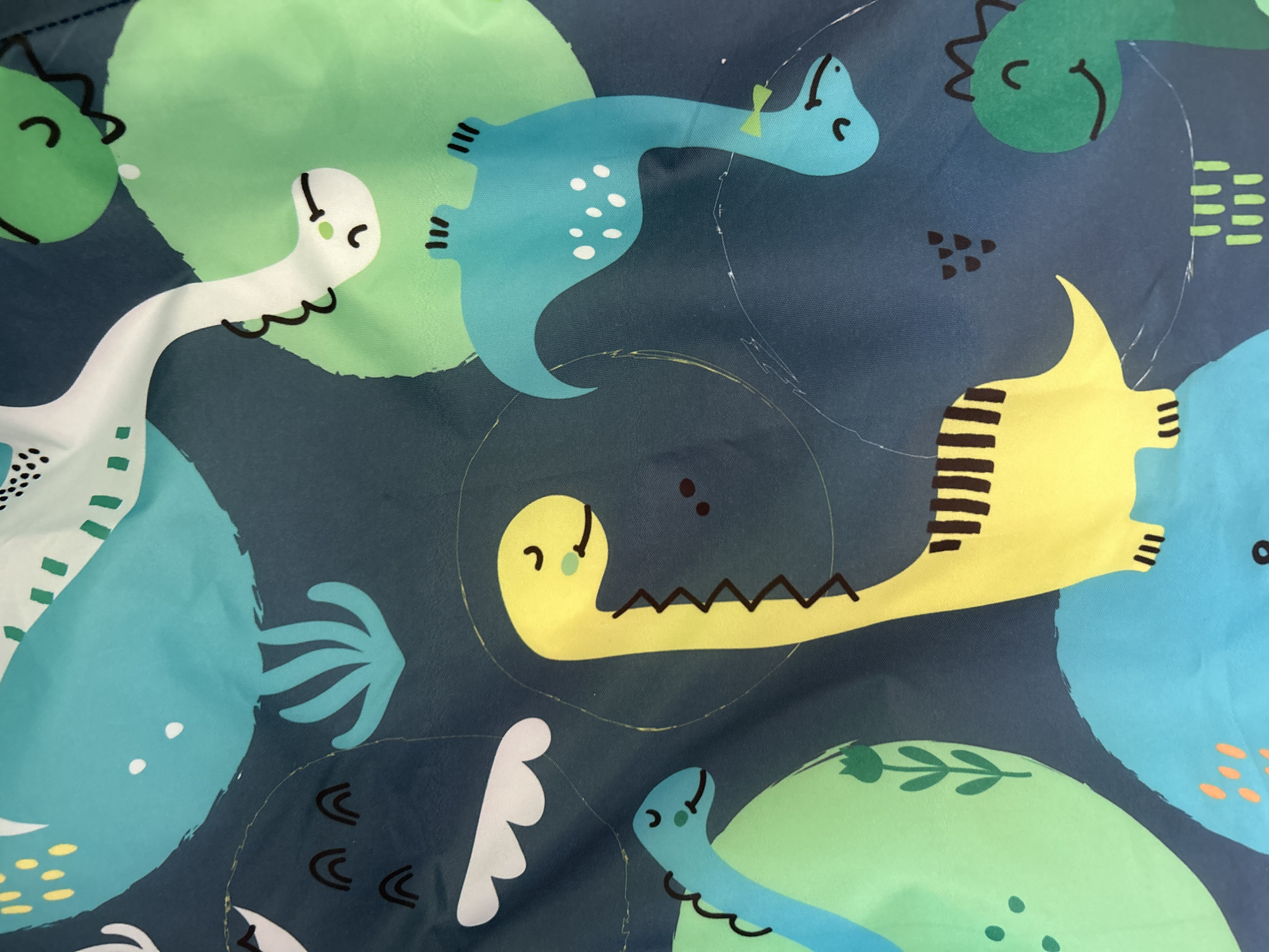 The image is a close-up, overhead shot of a fabric with a repeating pattern of cartoon dinosaurs. The dinosaurs are depicted in various colors including yellow, green, and blue, and are stylized with simple features like eyes and mouths. There are also abstract shapes and plant-like motifs scattered across the fabric. The background is a dark blue, giving the impression of a night sky or a deep ocean. The fabric appears to be a type of polyester or nylon, with a slightly shiny finish. There are visible wrinkles and creases in the fabric, suggesting it is either folded or has been laid out. No specific location in Khimki, Russia, or any other context is provided or discernible from the image itself.