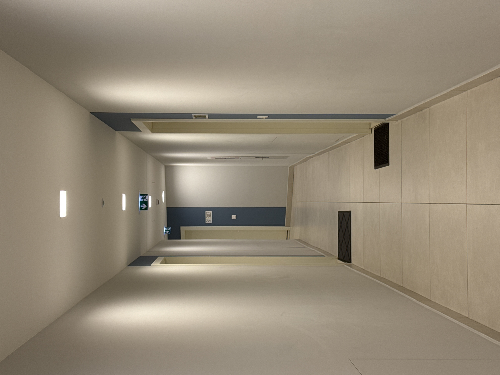 The image shows an indoor hallway with a light color scheme and fluorescent lighting. The walls are a light cream or off-white color, and the floor appears to be a similar shade of tile. The hallway stretches into the distance, with doors visible along the walls. There are illuminated exit signs present, as well as numbers visible on one of the doors. There are also dark, rectangular vents installed in the upper parts of the walls near the ceiling. The location in Belgrade, Serbia, is not discernible from the image.