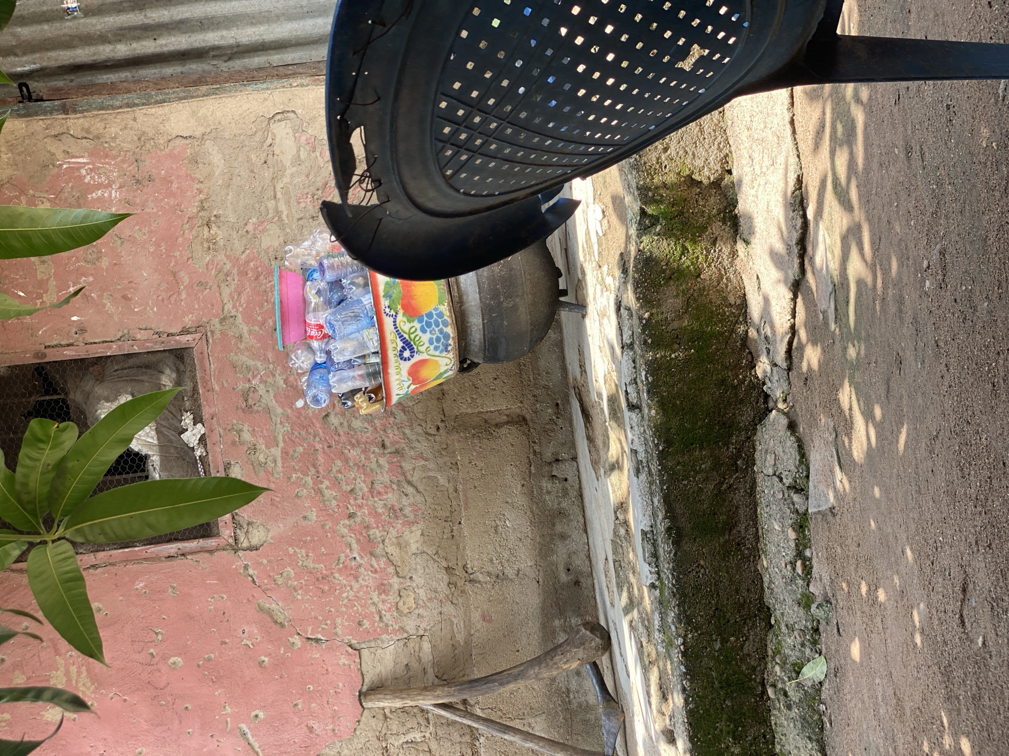 The image depicts an outdoor scene, likely in a residential area of Daniel, Nigeria. In the foreground, a dark plastic chair is partially visible, facing away from the viewer and angled to the right. Behind the chair, a collection of plastic water bottles is gathered in a large, decorative bowl. This bowl, featuring images of fruits like apples and grapes, sits atop a black, cast-iron pot with three legs. The pot is positioned on a raised concrete ledge, which also holds two wooden farming tools, leaning against a textured, pinkish-red wall. The wall is weathered and shows signs of cracking and peeling paint. To the right of the wall, a section of corrugated metal siding is visible. The ground in front of the ledge is made of reddish-brown dirt, with dappled sunlight creating shadows. Overhanging the scene from the top left are several large, green mango leaves. The overall impression is one of a functional, perhaps somewhat rustic, outdoor living or workspace.