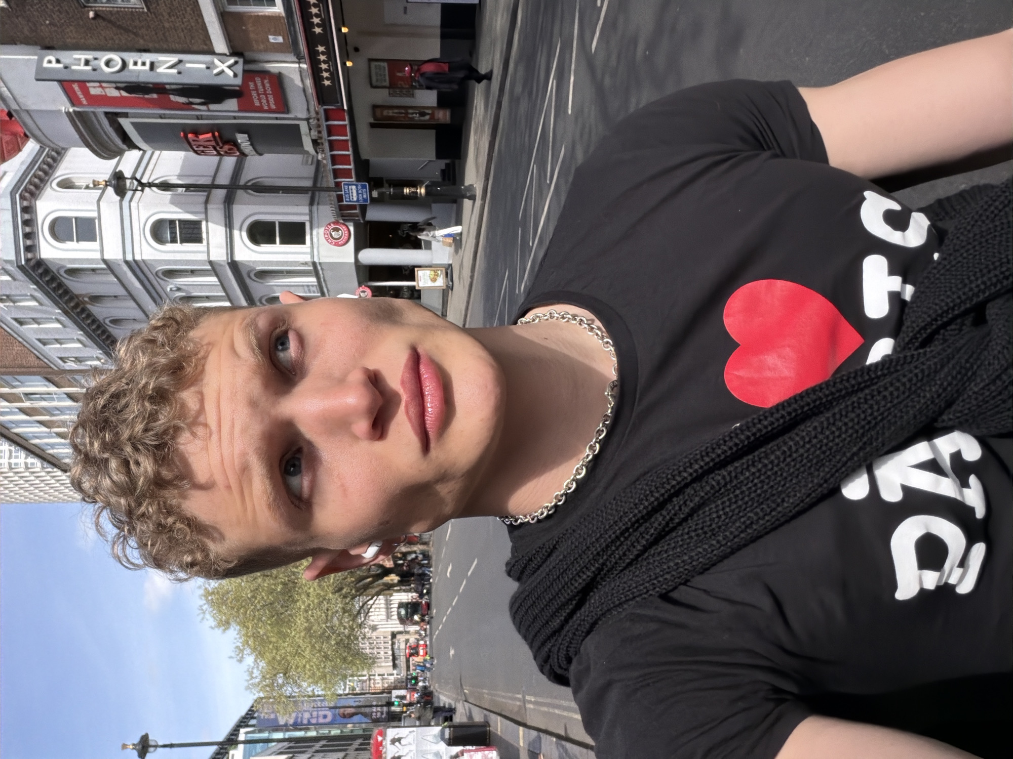 Here is a description of the image:
A young man is centered in the foreground, facing the camera with a slight upward tilt, wearing a black t-shirt with a red heart and white lettering. A black, knitted scarf is draped around his neck. The man is wearing a silver chain necklace.
In the background, the Phoenix Theatre is visible. Street-level activity includes pedestrians and vehicles. Trees are visible above buildings. The sky is visible in the upper right corner.
The location is in the city of London, United Kingdom.