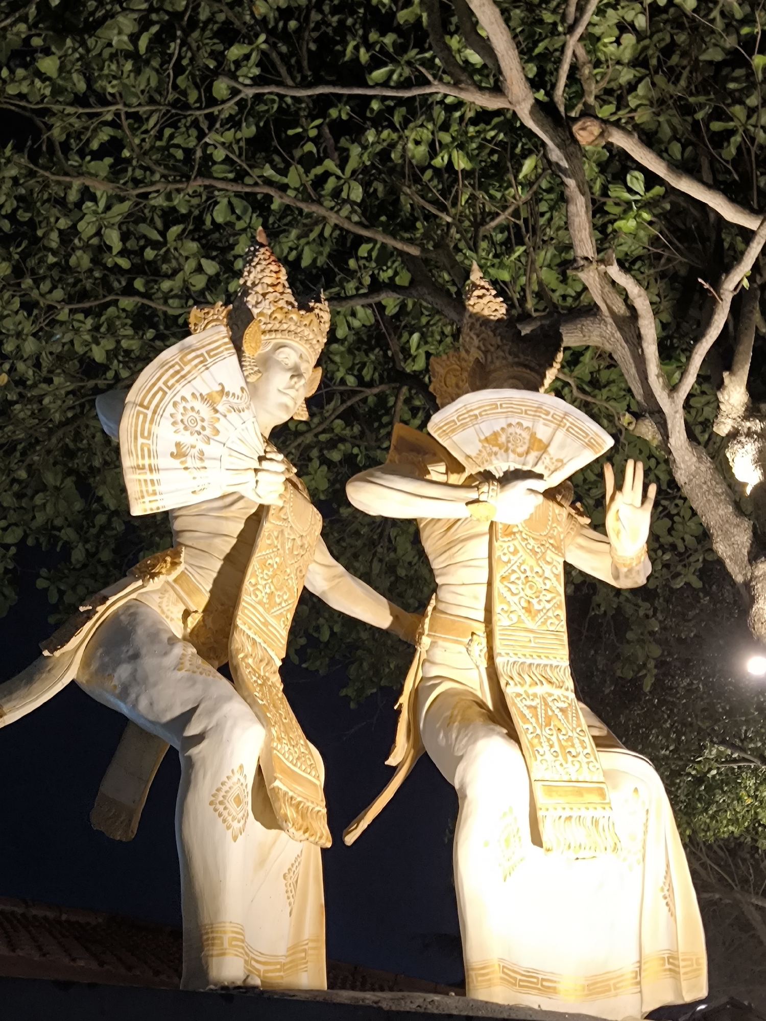 Two painted statues in traditional Balinese dress are posed in front of a tree with foliage, likely in North Kuta, Indonesia. Each statue holds a fan obscuring part of its face and is adorned with a patterned sash and crown. The statues are illuminated and appear to be outdoors at night.