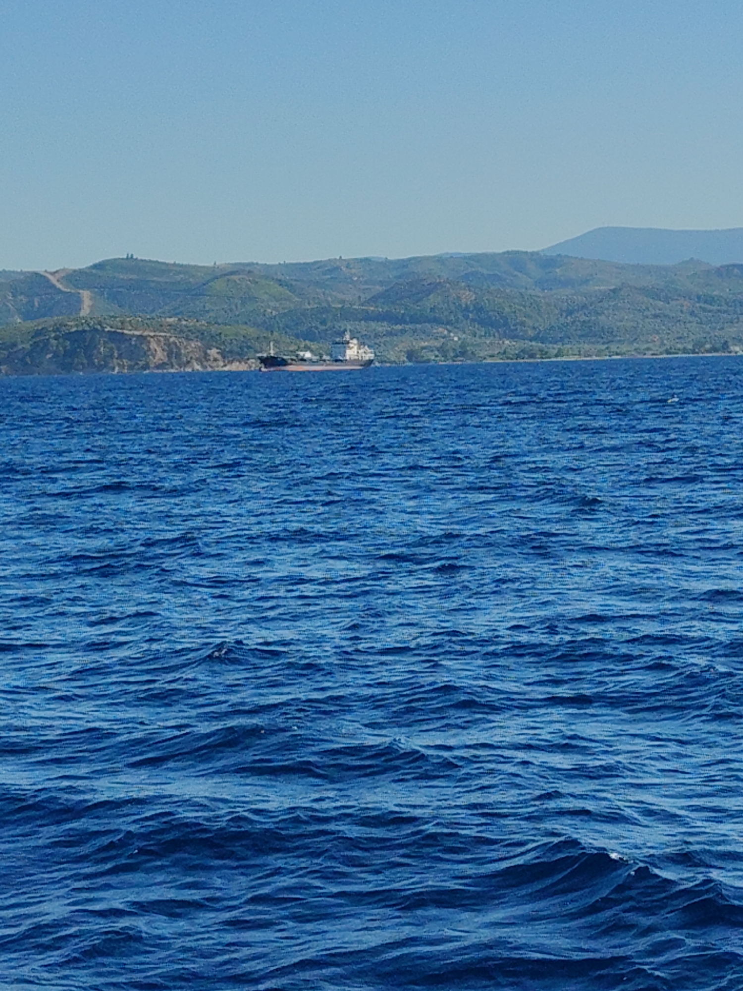 A wide shot captures a calm blue sea under a clear sky with distant green hills. In the middle ground, a large cargo ship is visible near the coastline of Paleokastro, Greece. The ship appears to be anchored or moving slowly. The water is rippled, suggesting a gentle breeze. The landscape features rolling hills covered in vegetation, with a few dirt paths winding up their slopes. The overall scene is serene and depicts a typical coastal view.