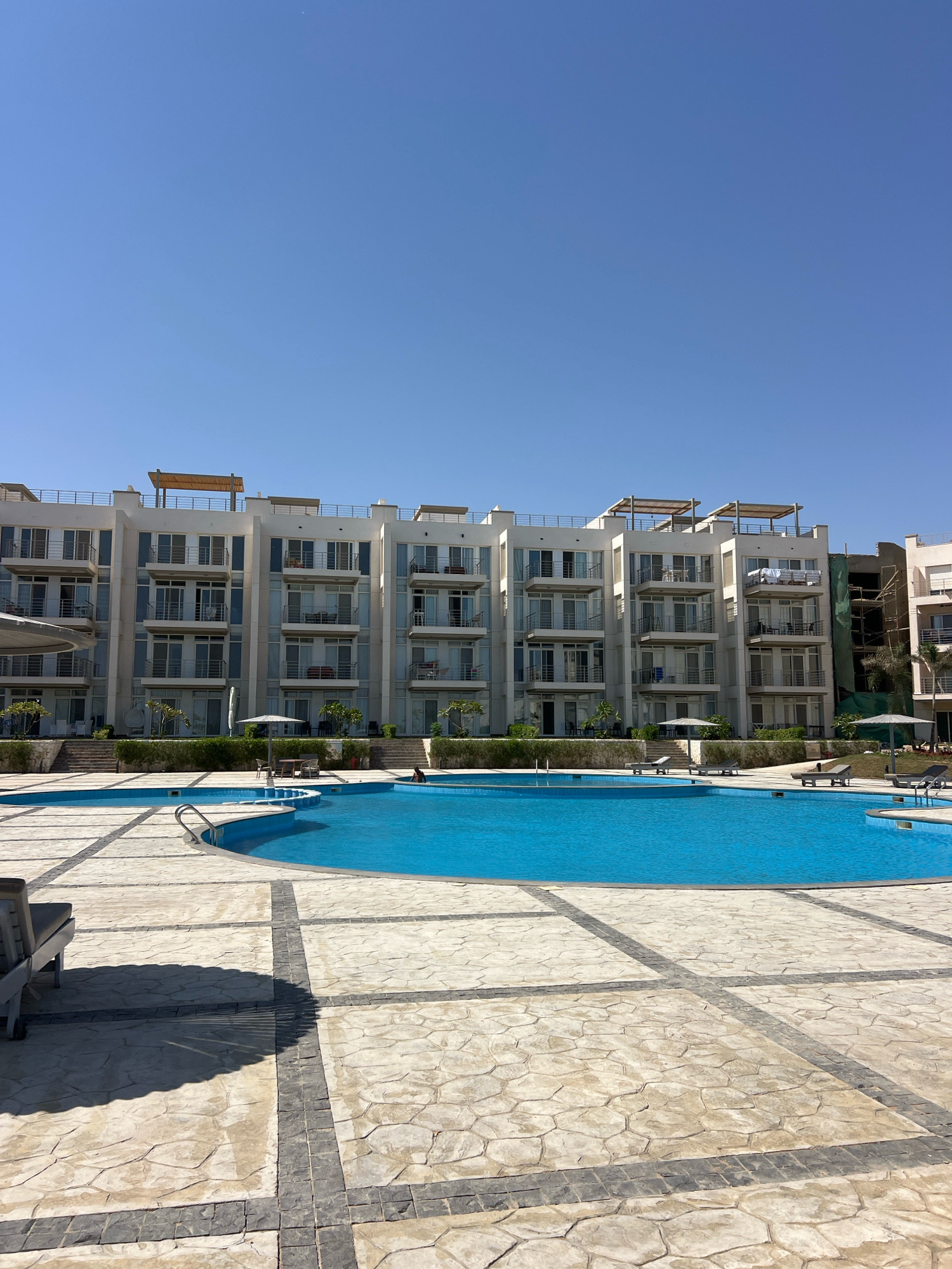 The image captures a bright, sunny day at a resort in Sharm El Sheikh, Egypt, featuring a large, vibrant blue swimming pool in the foreground. The pool has a curved shape with metal railings. The pool area is paved with large, light-colored rectangular tiles bordered by darker square tiles, creating a geometric pattern. Several empty lounge chairs are scattered around the pool.
Behind the pool, a multi-story white building with numerous balconies and large glass doors stretches across the frame. The building appears to be residential or hotel accommodations. Some balconies have furniture, and one has laundry hanging out to dry.
To the right of the main building, another building is partially visible, undergoing construction with scaffolding and green netting. Lush green landscaping with trees and shrubs is visible between the pool area and the buildings. The sky is a clear, cloudless blue. One person is visible in the pool, swimming. The overall scene depicts a well-maintained resort area ideal for relaxation and recreation.
