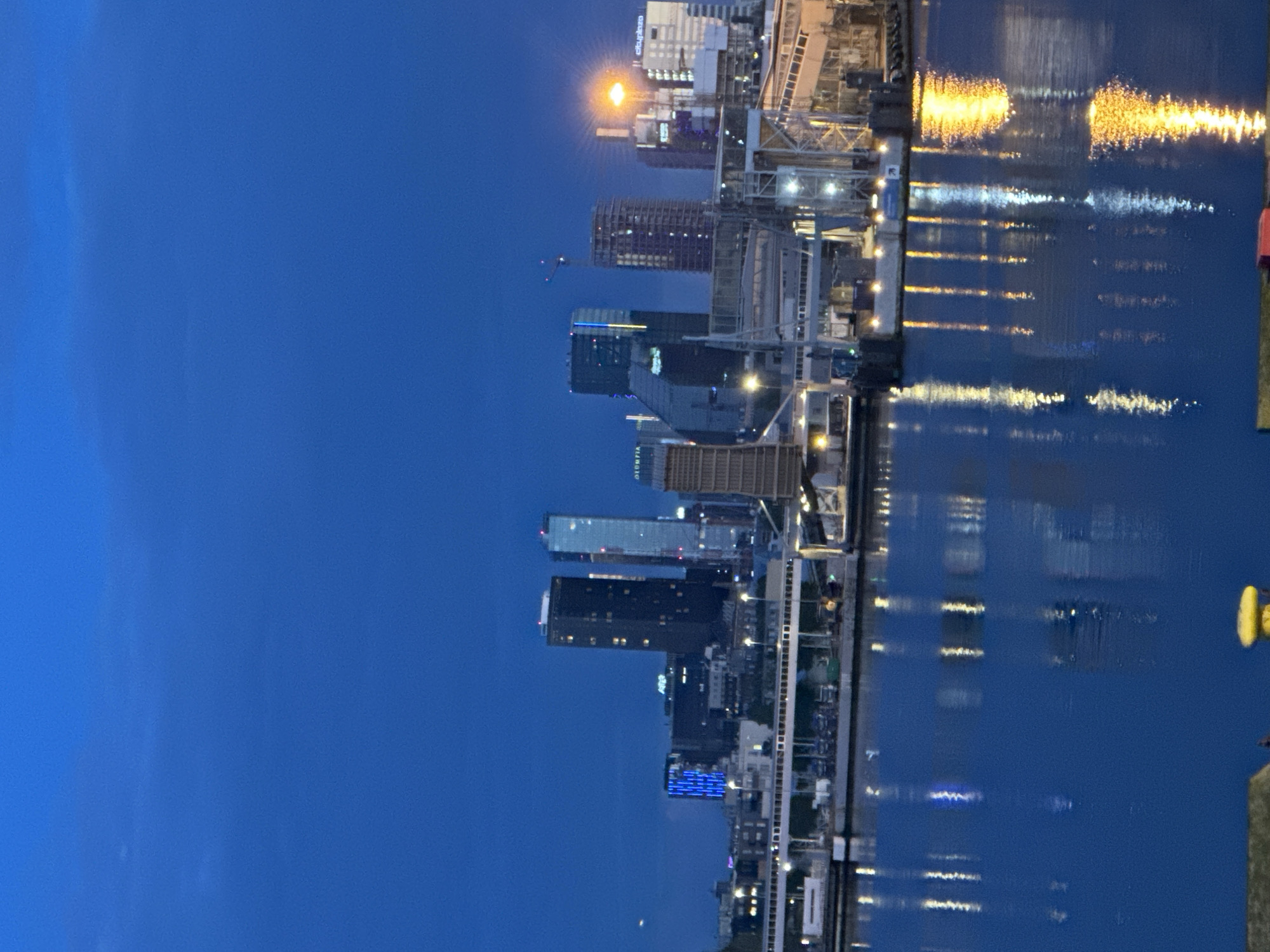 An aerial shot shows the Port of Tallinn in Estonia at dusk. The deep blue sky is visible, with reflections of city lights shimmering on the water. The cityscape features several modern, tall buildings, including a dark rectangular skyscraper with illuminated windows and a glass-fronted building with blue lights. The port itself has various structures, including walkways, cranes, and cargo containers. The water is calm, creating clear reflections of the lights from the buildings and the port.
