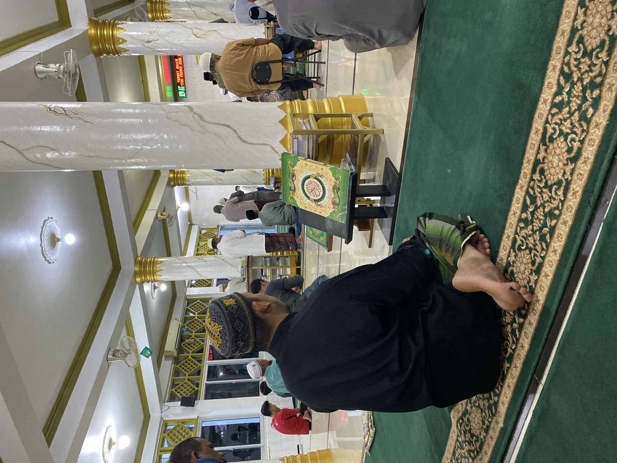 The image depicts the interior of a mosque, likely in Lhokseumawe, Indonesia, during prayer time. Several men are visible, some seated on a green carpeted floor with ornate golden patterns, while others are standing or in prayer. The mosque features white marble-patterned columns with gold accents and a white ceiling with decorative light fixtures and ceiling fans. In the foreground, a man wearing a black shirt and a patterned Kufi hat is seated, facing away from the camera. Next to him, on the floor, is a green Quran on a black stand. Behind him, other men are engaged in prayer or awaiting prayer. A digital display showing prayer times is visible in the background. The overall scene suggests a communal religious gathering.
