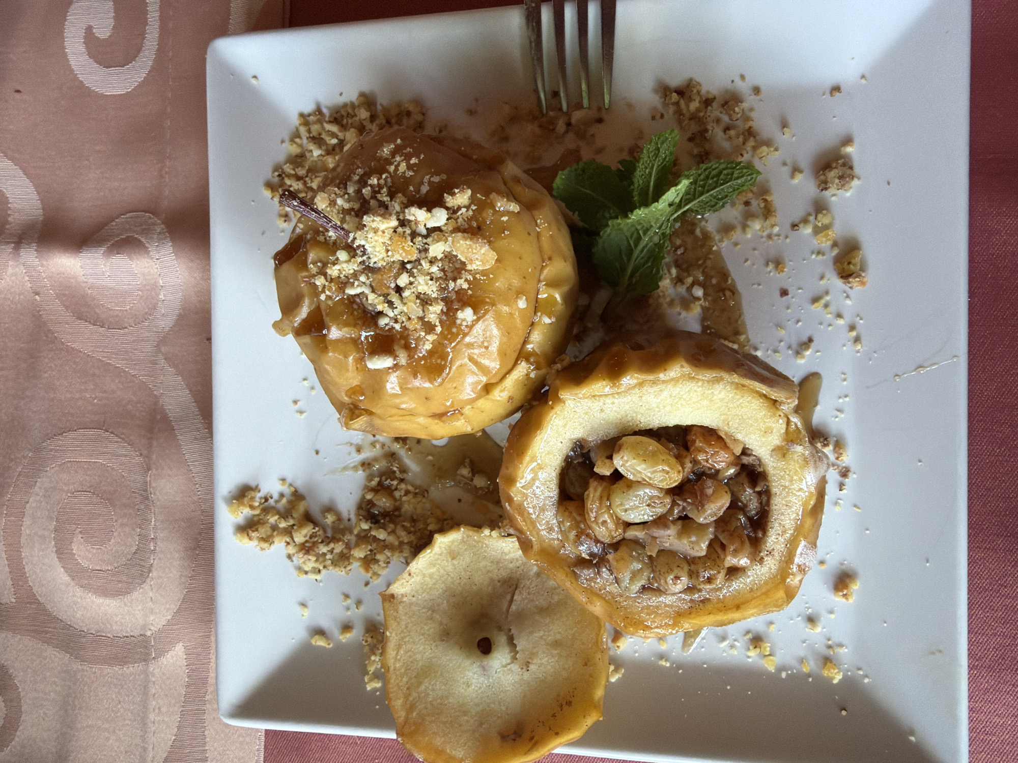 A top-down shot captures a square, white plate featuring a dessert, possibly served in Pleven, Bulgaria. Two baked apples dominate the scene; one is whole, glazed, and sprinkled with crushed nuts, while the other is halved, revealing a filling of nuts and raisins. A separate slice of baked apple sits near the halved apple. A sprig of fresh mint serves as garnish. A fork rests behind the whole apple. Crushed nuts are scattered around the apples on the plate. The plate is set on a patterned tablecloth with tones of peach and pink.