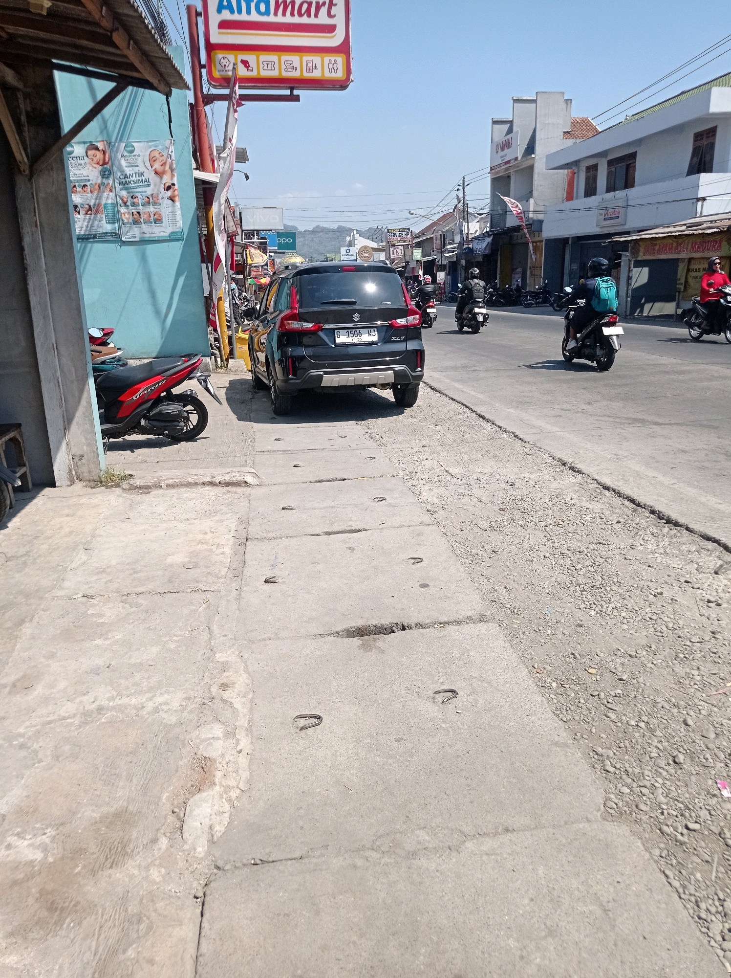 This is a street scene in Karangtuang, Indonesia, on a bright, sunny day. The perspective is from a sidewalk looking towards a road. On the left, a blue-walled building with a wooden overhang is visible, displaying posters advertising beauty products. A red and black motorcycle is parked near the building. A black car with license plate "G 1506 HJ" is the primary focus in the middle ground, partially obscuring the road and sidewalk. Several other motorcycles, some with riders, are on the road, moving away from the viewer. The road is paved and appears to be a busy street with buildings lining both sides. The sidewalk is made of concrete slabs, showing some wear and tear. The environment suggests a commercial area with small businesses.
