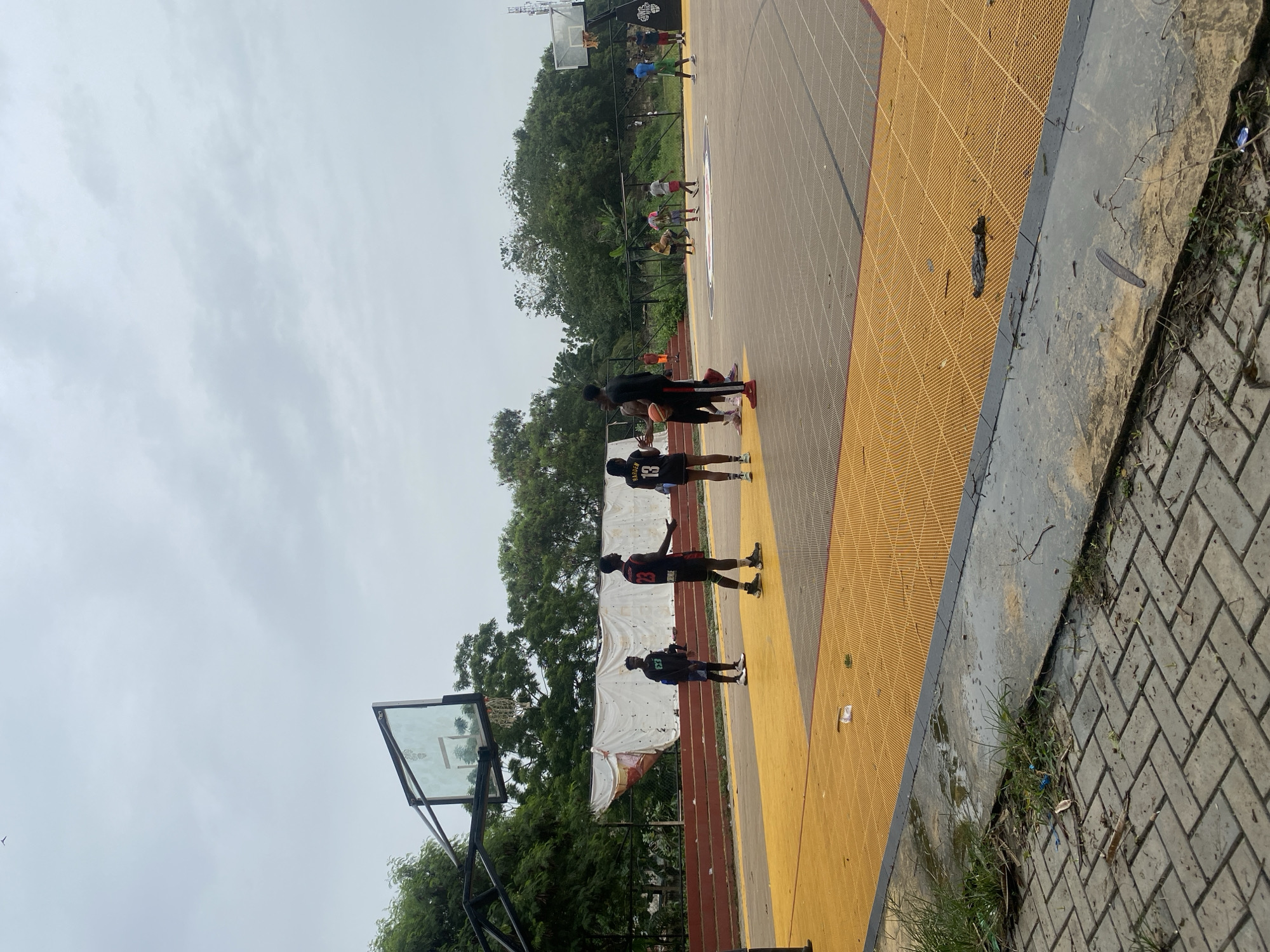 The image depicts an outdoor basketball court, likely in Lagos, Nigeria, given the style of the court and general environment. The court has a yellow playing surface with a grey border. There are several people on the court, including a group of four young men standing near one hoop and other individuals playing further down the court. The men are wearing athletic clothing. The environment consists of greenery like trees, and a cloudy sky. The court appears to be adjacent to a paved area and some vegetation. There is a basketball hoop and backboard at the bottom of the image.