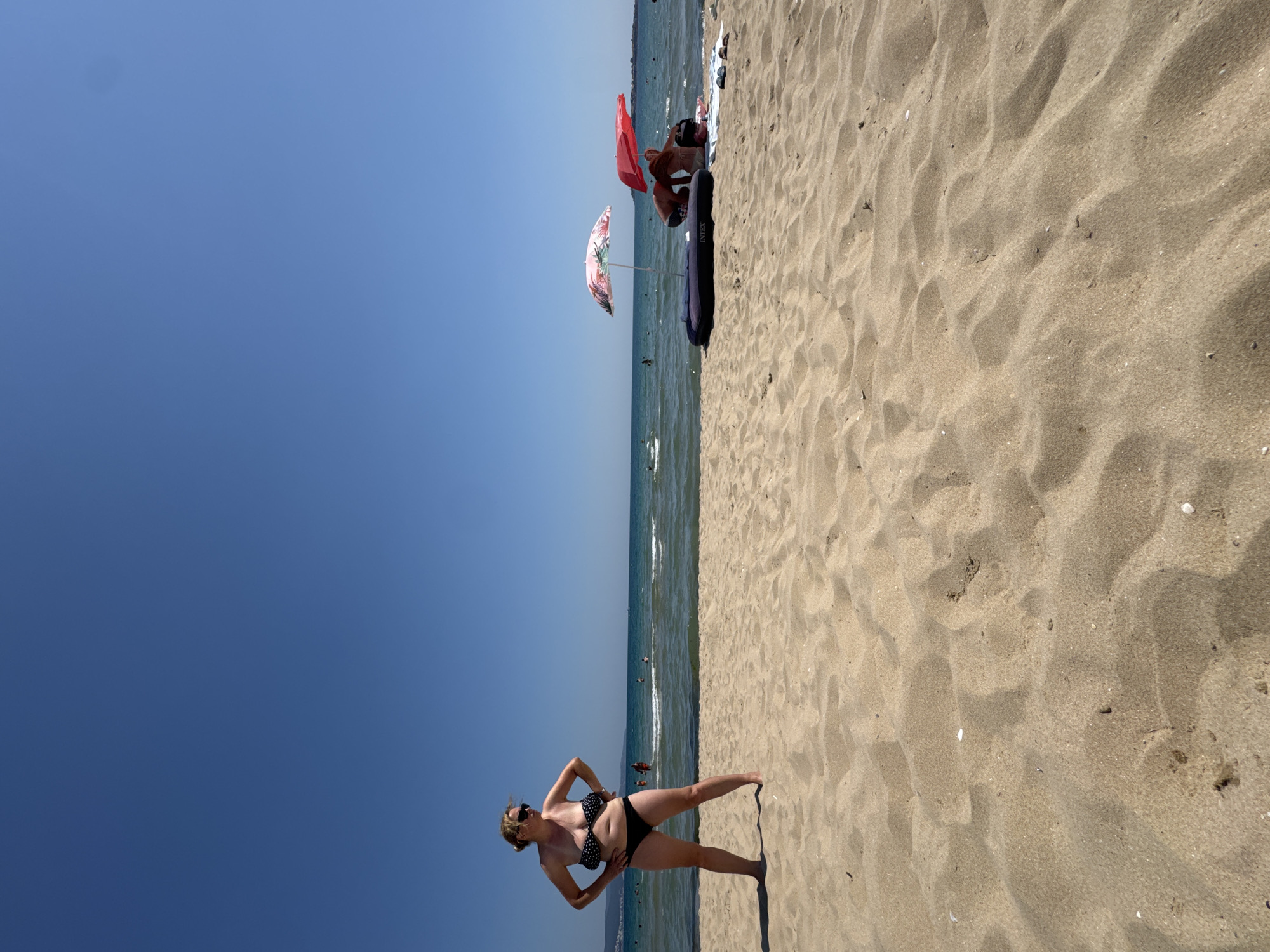 A wide-angle, eye-level shot captures a sunny day at a sandy beach in Nesebar, Bulgaria. The foreground is dominated by rippled sand dunes, leading to the shoreline. In the mid-ground, a woman stands with her hands on her hips, wearing a polka-dot bikini and sunglasses. She is facing the water, with the sea and a clear blue sky behind her. Further to the right, two people are seated under umbrellas near an inflatable raft on the beach. The sea is calm with small waves, and several other people are visible in the water. The coastline with distant buildings is visible on the horizon. The scene depicts a typical beach day with leisure activities.