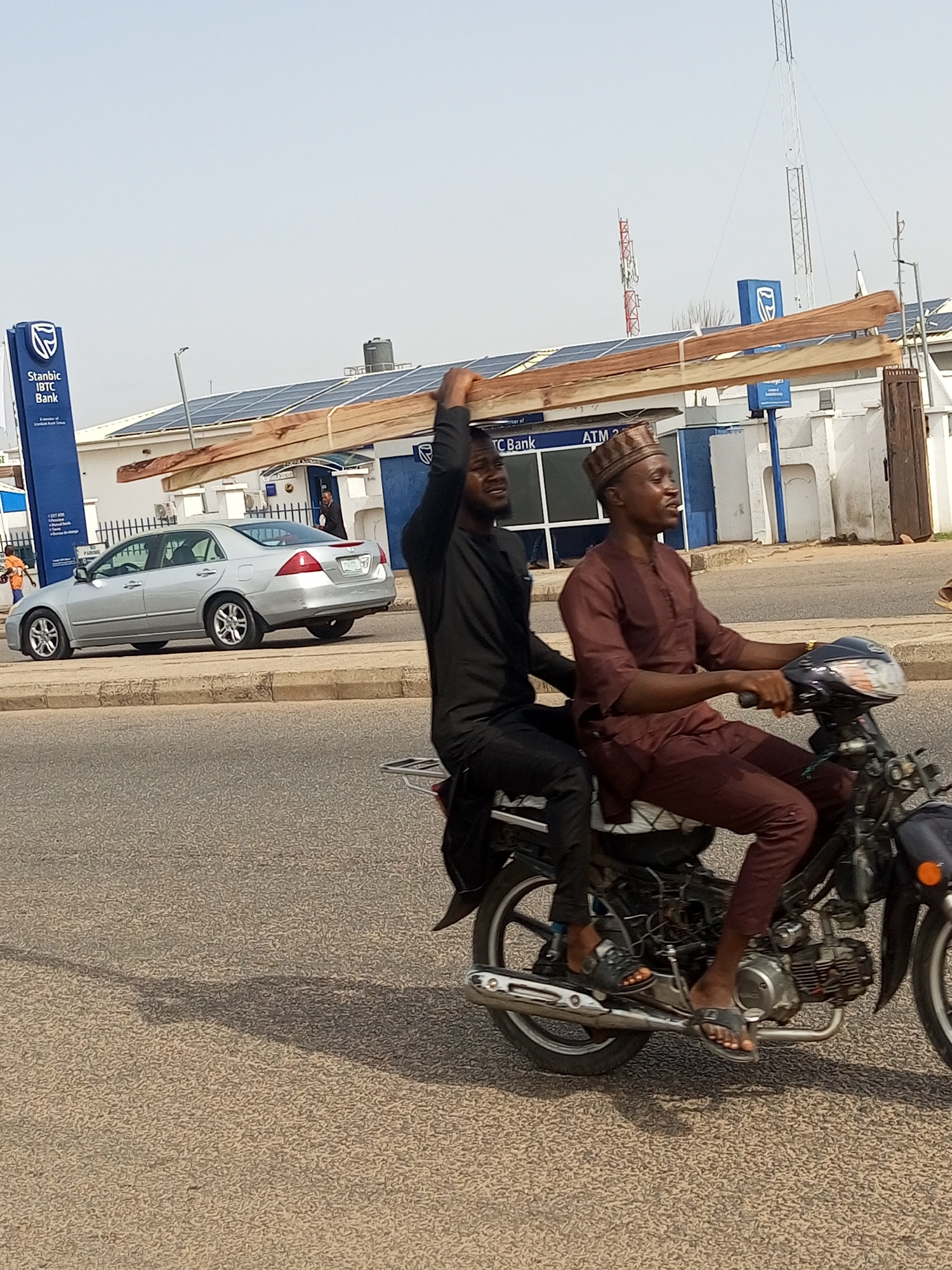 Two men are riding on a motorcycle, transporting long wooden planks. The man in front is wearing a brown traditional outfit and a patterned cap, and is operating the motorcycle. The man behind him, wearing a black outfit, is holding the wooden planks with one hand, and appears to be looking upwards. The motorcycle is traveling on a paved road. In the background, there is a silver car, a building with "Stanbic IBTC Bank" signage, solar panels on the roof of the building, and telecommunication towers. The environment suggests an urban setting.