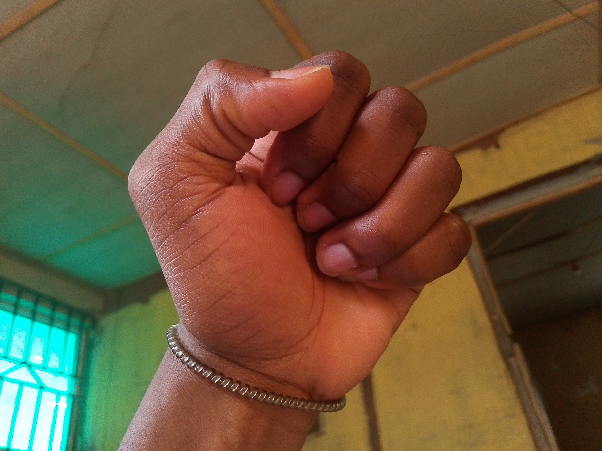 A close-up shot shows a brown-skinned hand clenched into a fist, with a simple, coiled metal bracelet around the wrist. The background reveals portions of a room with green and yellow walls. A window with a teal-painted frame and grid-like design is visible. The location is Ipetumodu, Nigeria. The scene lacks any identifiable people or subjects besides the arm and hand.