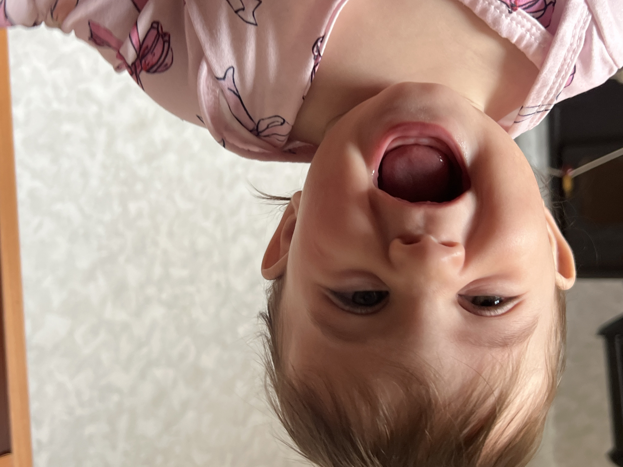 A baby is presented upside down, wearing a pink garment patterned with bows. The baby's mouth is open wide. The backdrop is a wall covered with a repeating pattern. The location is potentially Elena, Bulgaria, however no confirmatory evidence can be provided.