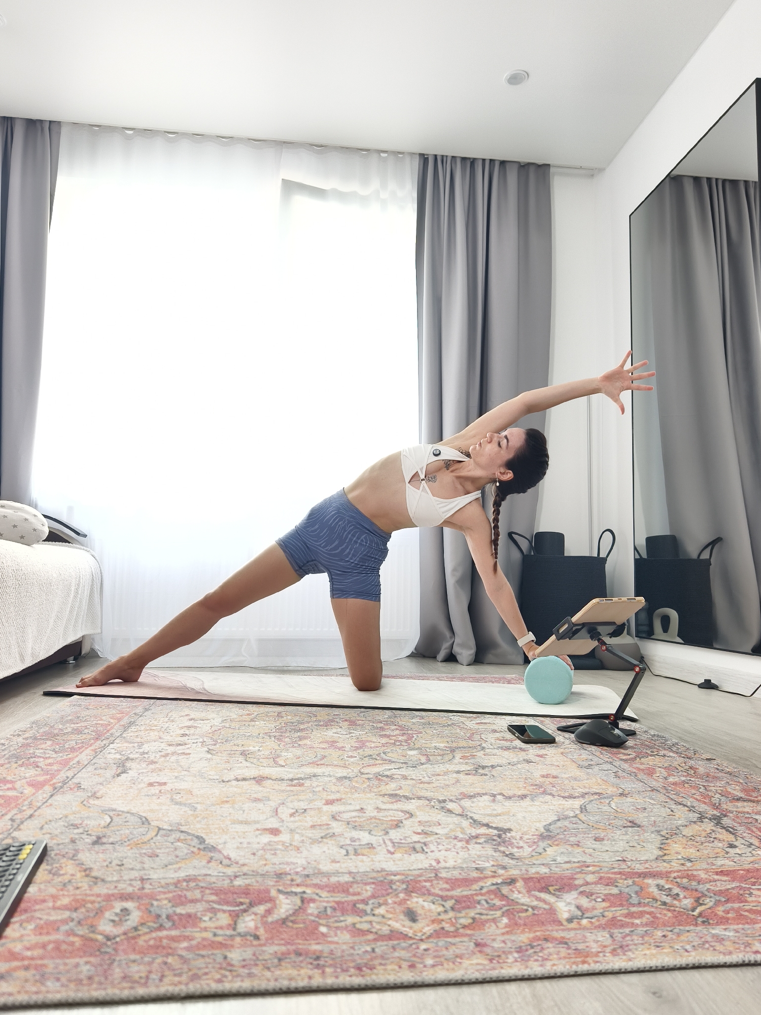 A woman is performing a yoga pose on a mat in a well-lit room. She is kneeling on her right knee with her left leg extended to the side. Her torso is bent to the left, with her left arm extended upwards and her right arm reaching towards the floor. She is wearing a white sports bra and blue patterned shorts. In front of her, there is a tablet on a stand, a light blue exercise ball, a smartphone, and a computer mouse. The room has grey curtains, a mirror on the right wall, and a rug with a red and beige pattern on the floor. The overall setting appears to be a home interior.