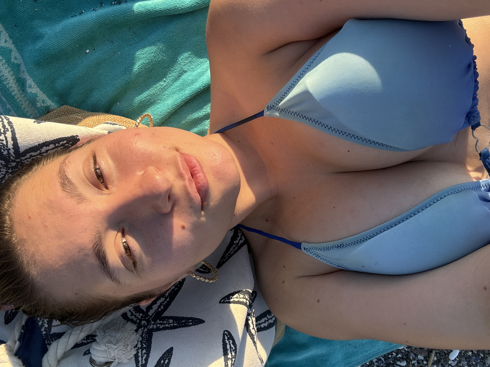 The media file shows a close-up, overhead shot of a young woman lying on a beach. She is wearing a light blue bikini and is positioned on a striped towel, which is laid out on pebbles. The woman's face is partially obscured by shadow, but she appears to be looking up. She has a gold hoop earring in her left ear. The background consists of a turquoise towel, suggesting a beach or poolside setting. The lighting indicates it is daytime, likely sunny, with the sun casting a warm glow on the woman's skin. The scene evokes a relaxed and leisurely mood, typical of a day at the beach. No text is visible in the image. The location context is Sozopol, Bulgaria.