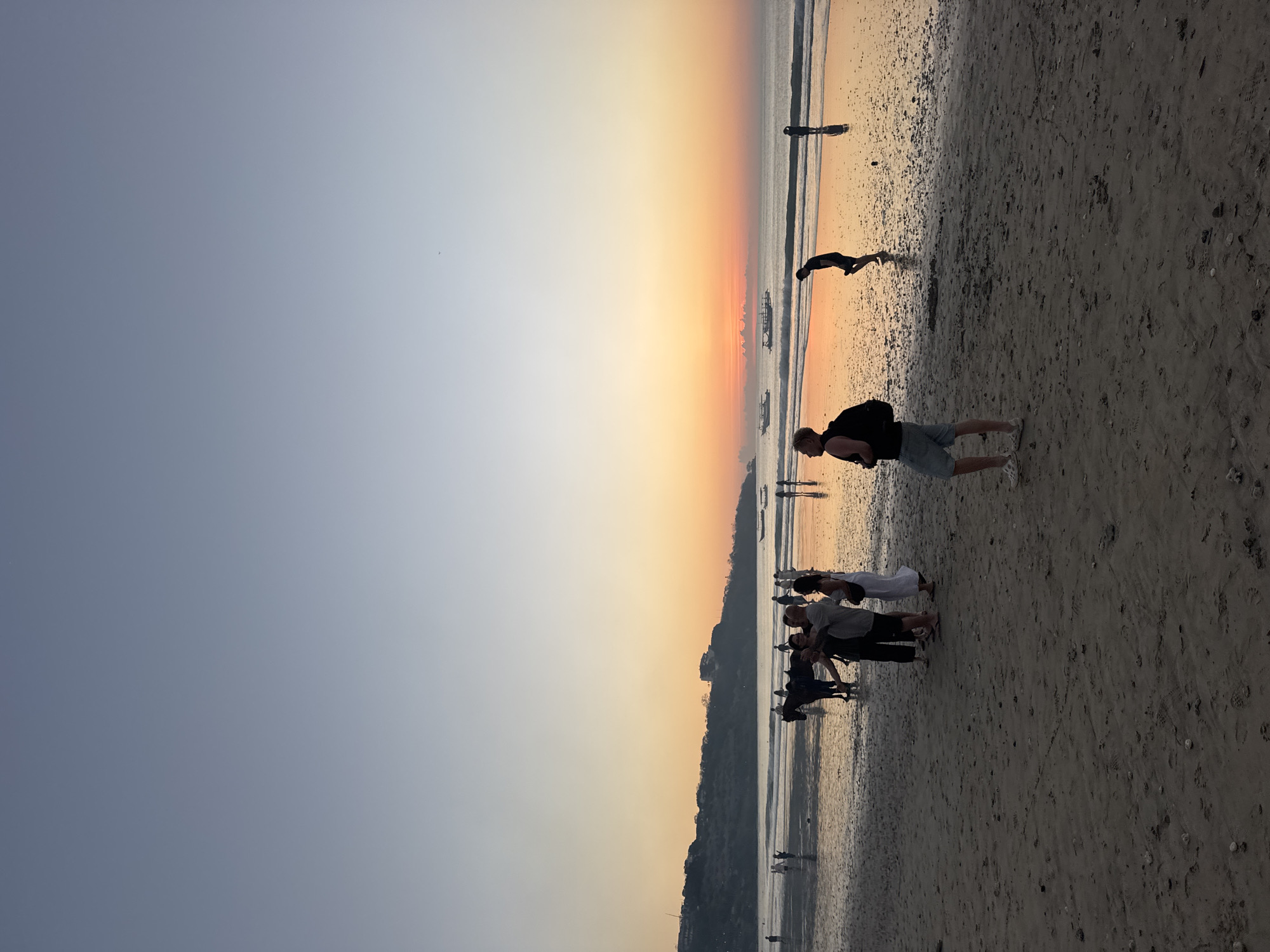 The image depicts a beach scene at sunset in Kuta Selatan, Indonesia. The sky transitions from a clear, pale blue at the top to a warm orange and yellow gradient near the horizon, indicating the setting sun. The ocean is calm with gentle waves lapping at the shore. Several boats are visible in the distance on the water.
On the wet sand of the beach, there are numerous people. In the foreground, a man with a backpack stands facing away from the camera, wearing a black t-shirt and denim shorts. To his left, a group of people are gathered, with one individual taking a photo of the others. Among this group, a woman in a white dress is visible. Further back, closer to the water's edge, are more individuals and a horse being ridden. The sand itself is damp and reflective, showing the colors of the sunset. In the background, a hilly coastline with some buildings can be seen. The overall atmosphere is one of a peaceful evening at the beach.