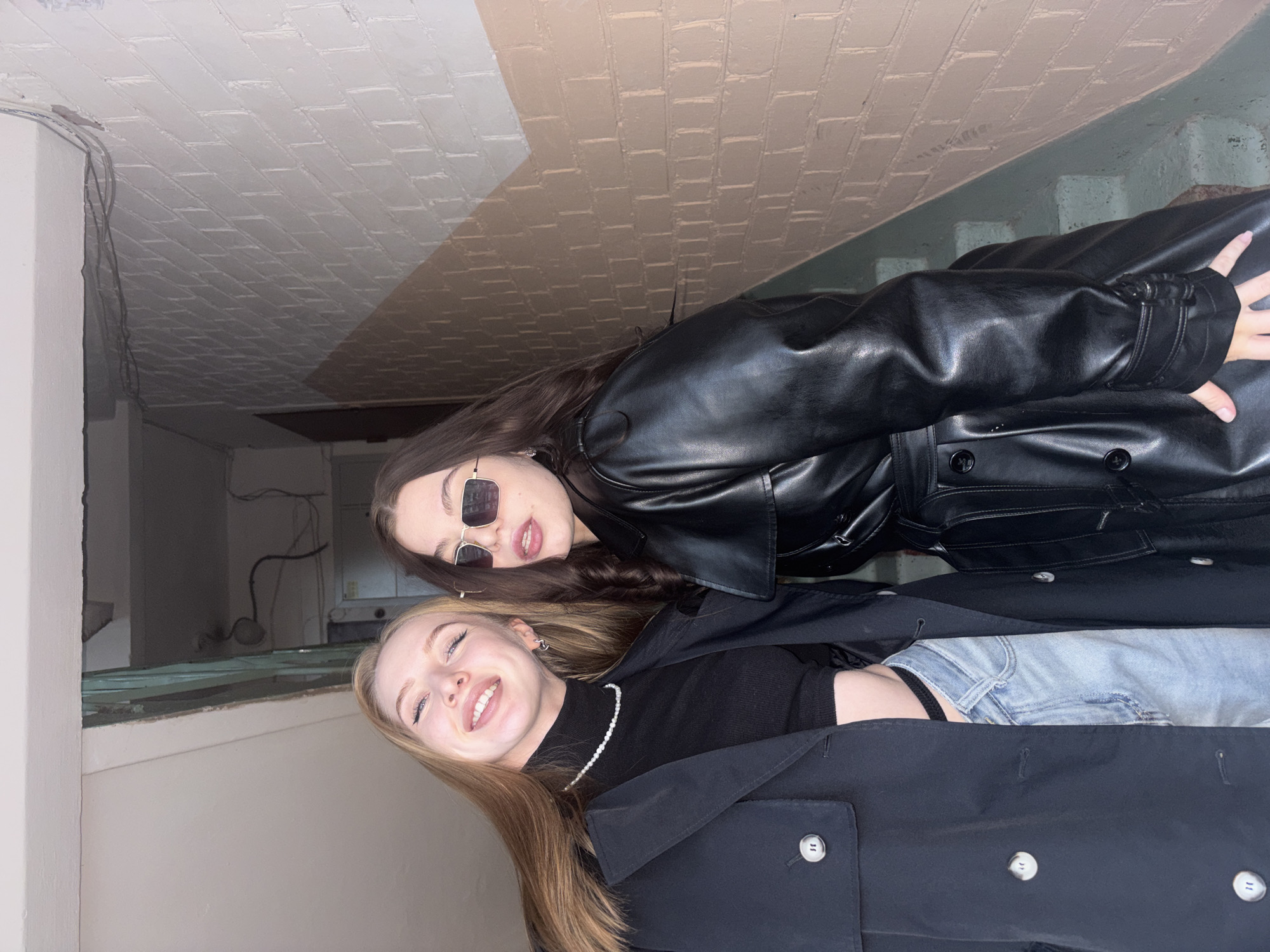 Two young women pose in an indoor space. One is blonde with long hair, wearing a black turtleneck, light-wash jeans, a pearl necklace, and a dark trench coat. She is smiling. The other woman has dark hair and is wearing sunglasses and a black leather jacket. They appear to be lying down. The environment includes brick walls, possibly in a building located in Perm, Russia, judging by architectural features.