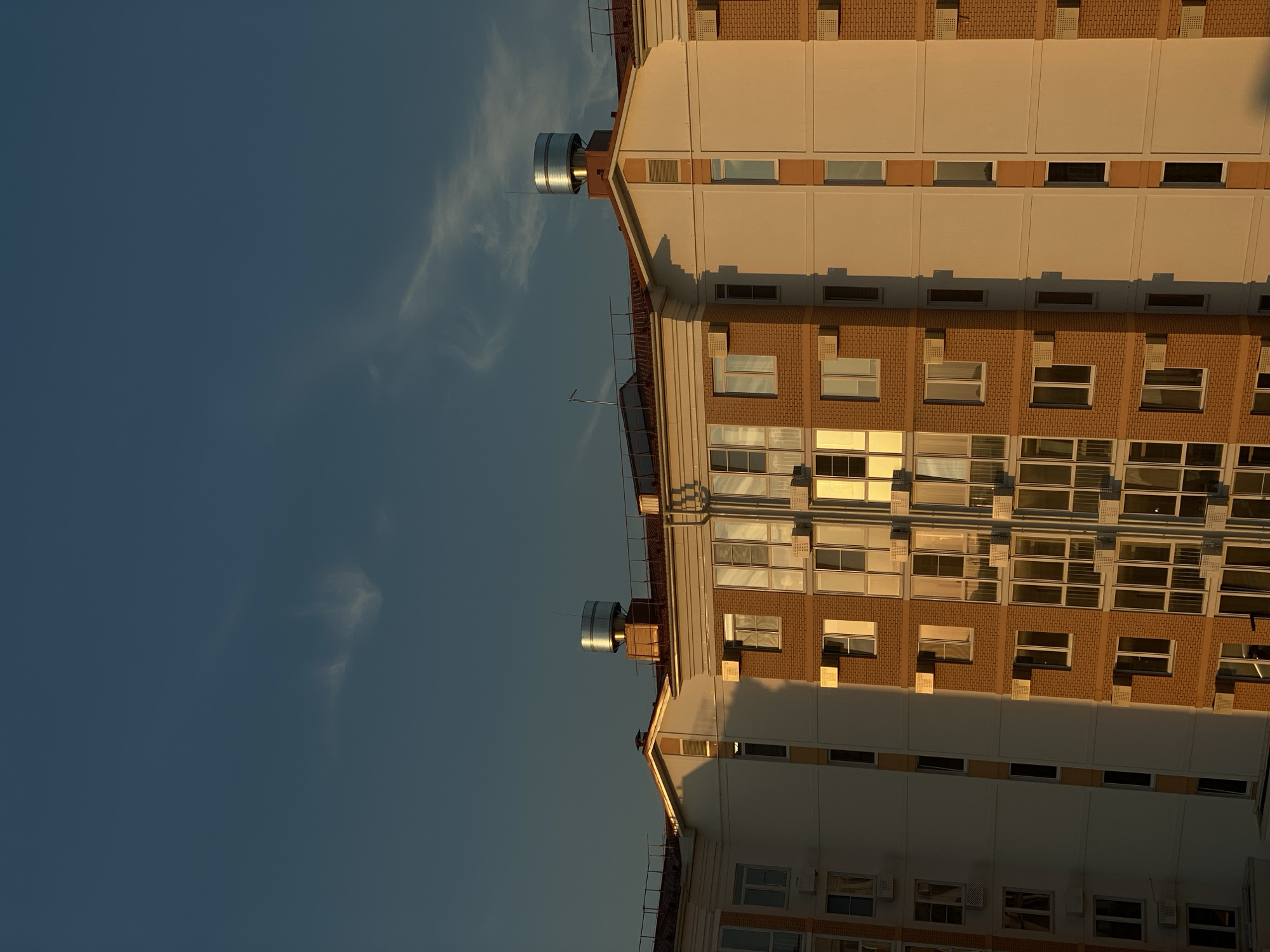 The image shows the upper floors of a modern apartment building bathed in the warm, golden light of a setting or rising sun. The building features a combination of white and light beige facades with sections of orange brickwork. Numerous windows, many with reflective glass, are visible, along with external air conditioning units mounted below some of them. The roofline is visible, with a prominent silver cylindrical structure, possibly a ventilation system or water tank, on one section. A few wispy clouds are present in the deep blue sky. The scene is devoid of any people or visible activity, presenting a static architectural view. The context provided suggests this location is in Stopyatyy, Russia, though the image itself does not contain any specific identifying landmarks to confirm this.