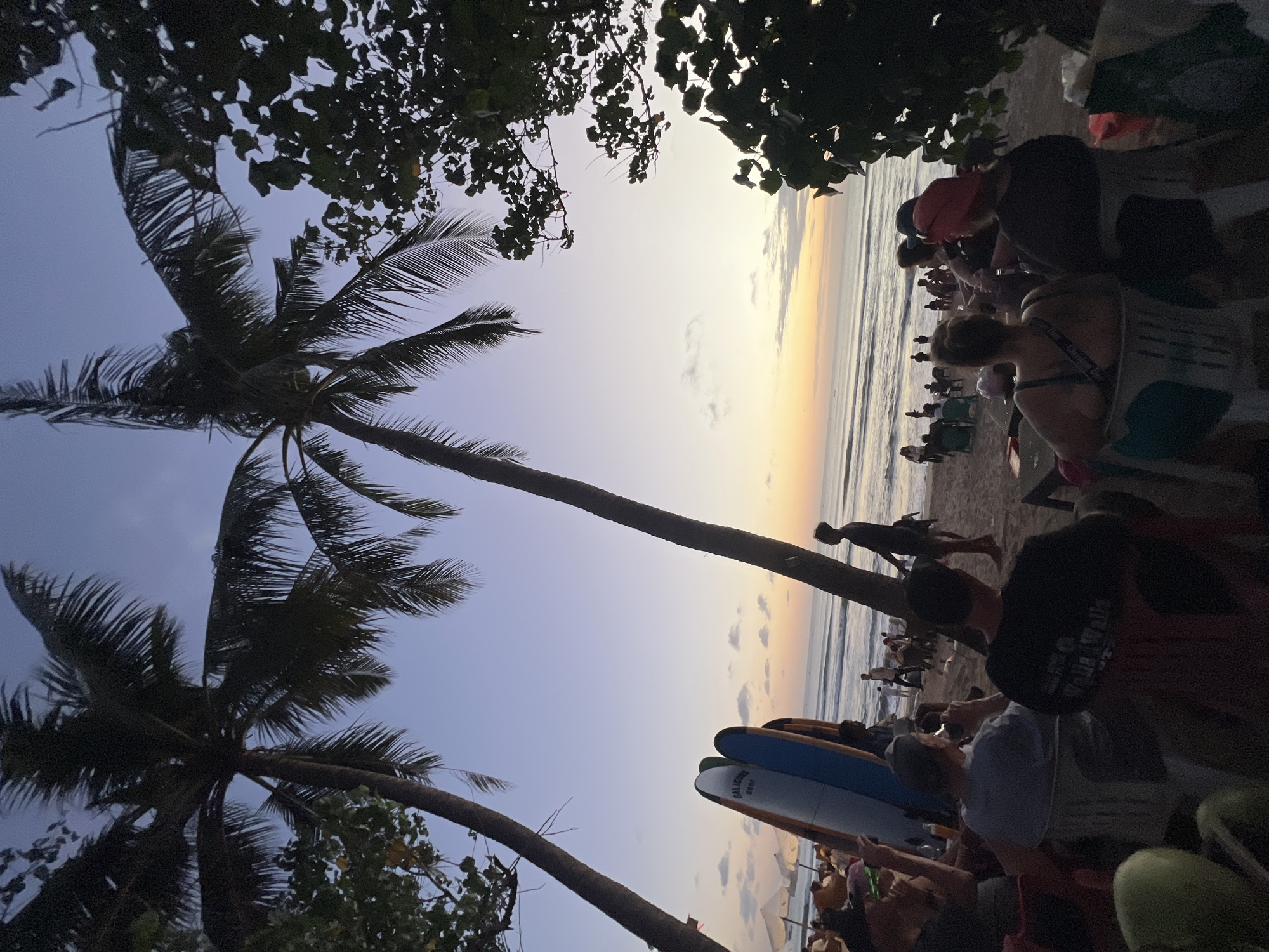 This image depicts a beach scene in Kuta, Indonesia, during twilight. The sky is a gradient of soft purples, blues, and oranges as the sun sets over the ocean, casting warm light on the water. Two prominent palm trees frame the view, their fronds silhouetted against the sky. In the foreground, several people are seated on chairs on the sand, facing the ocean. Some appear to be enjoying drinks and conversation, while others are looking towards the water. Behind them, a group of surfboards are propped upright. Further down the beach, more people are visible, some standing and walking near the water's edge. The overall atmosphere is relaxed and social, suggesting a popular spot for evening enjoyment.