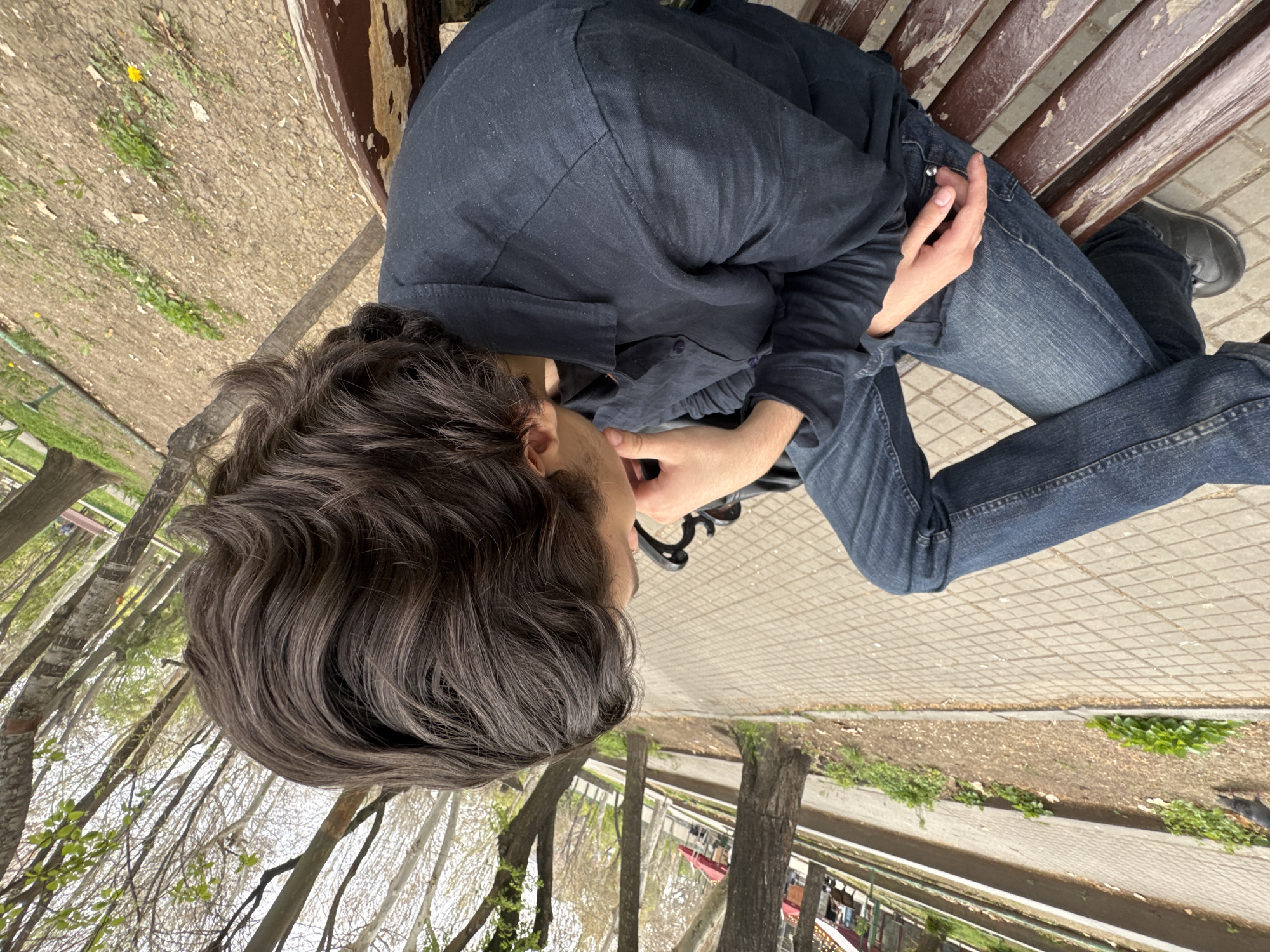 A young person with wavy brown hair is sitting on a brown wooden bench in a park. They are wearing a dark blue button-up shirt and blue jeans, with grey shoes visible on their right foot. The person is positioned at an angle, facing left, and is looking downwards. The background includes a paved path, grass, trees, and a red structure, possibly a train or carousel. The location appears to be in a park setting, but no specific location in Yerevan, Armenia can be identified from the image.