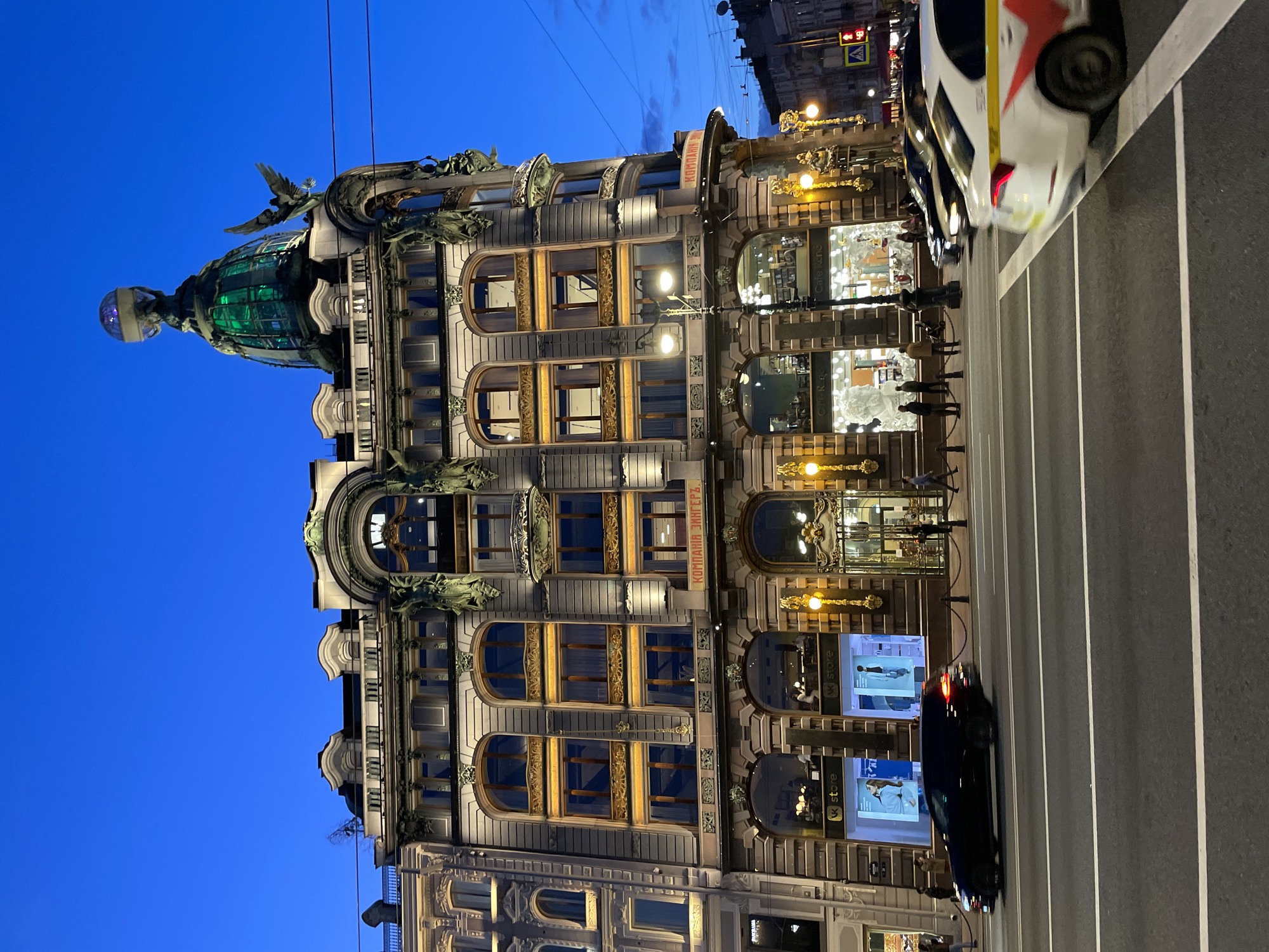 The image displays an evening or nighttime scene in Saint Petersburg, Russia, featuring the grand, ornate architecture of the Singer House (Dom Knigi). The sky is a deep, clear blue, characteristic of twilight or night, providing a stark contrast to the brightly illuminated building.
The multi-story building, an example of Art Nouveau architecture, dominates the left and center of the frame. Its facade is adorned with intricate classical details, decorative sculptures, and large arched windows. On the left, a distinctive green-domed tower rises, capped with a glass sphere and a large, elegant sculpture of a bird (likely an eagle) with outstretched wings. Many windows feature elaborate golden framework and decorative elements.
On the ground floor, several brightly lit storefronts are visible, displaying merchandise. The text "VK store" is clearly seen on multiple screens within these shops, and "Cafe Luno" or "Cafe Lumo" is partially visible on another. High on the building, the historical inscription "КОМПАНИЯ ЗИНГЕРЪ" (KOMPANIYA ZINGER) is prominent, confirming the building's identity.
In the foreground and to the right, a multi-lane street is depicted with visible road markings. Several vehicles are present, including a white car with red and yellow livery (suggesting a taxi) and a dark car. People are seen walking on the sidewalk in front of the building, appearing as dark figures against the illuminated shop windows. Streetlights provide additional illumination, and power lines traverse the sky overhead. A pedestrian crossing sign displaying a red light is also visible in the upper right. The scene conveys a sense of a lively urban environment after sunset.