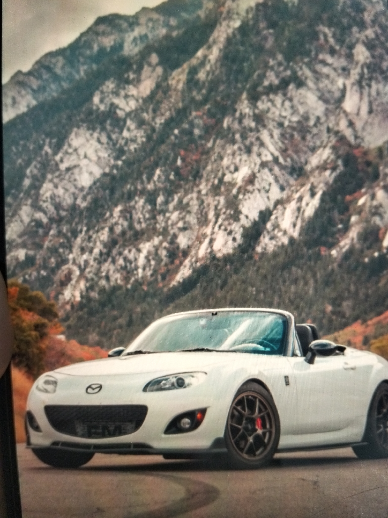 A white convertible Mazda Miata is parked on a road with a mountain range in the background. The mountains are rocky and covered with trees. The sky is cloudy. The car has black wheels with red brake calipers. There is a license plate on the front of the car that is partially visible. The car is in good condition and appears to be well-maintained. The location of Payagaboh, Indonesia is not depicted in this image.