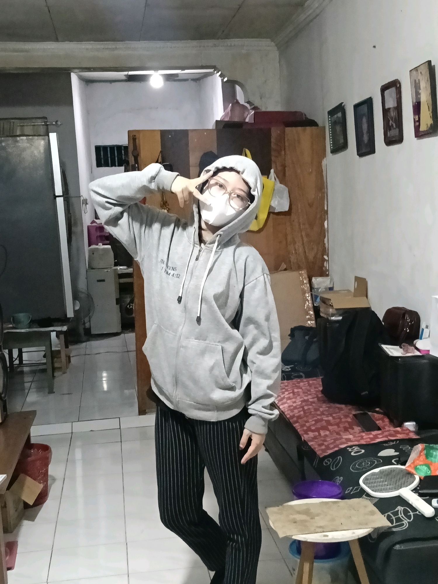 A person wearing a grey hooded sweatshirt and black and white striped pants stands in a room with tiled floors and concrete walls. The person is wearing a white mask and glasses and has their hood up. They are making a peace sign with their right hand. In the background, there is a refrigerator, a fan, a wooden cabinet, and several framed pictures on the wall. There is also a bed with a patterned blanket and various objects on it, including a white hand mirror with a black handle and a purple bucket. A small wooden table with a piece of cardboard on top is in the foreground. The lighting in the room is dim, with a single lightbulb visible on the ceiling. The scene appears to be an indoor residential space in Jatimulya, Indonesia.