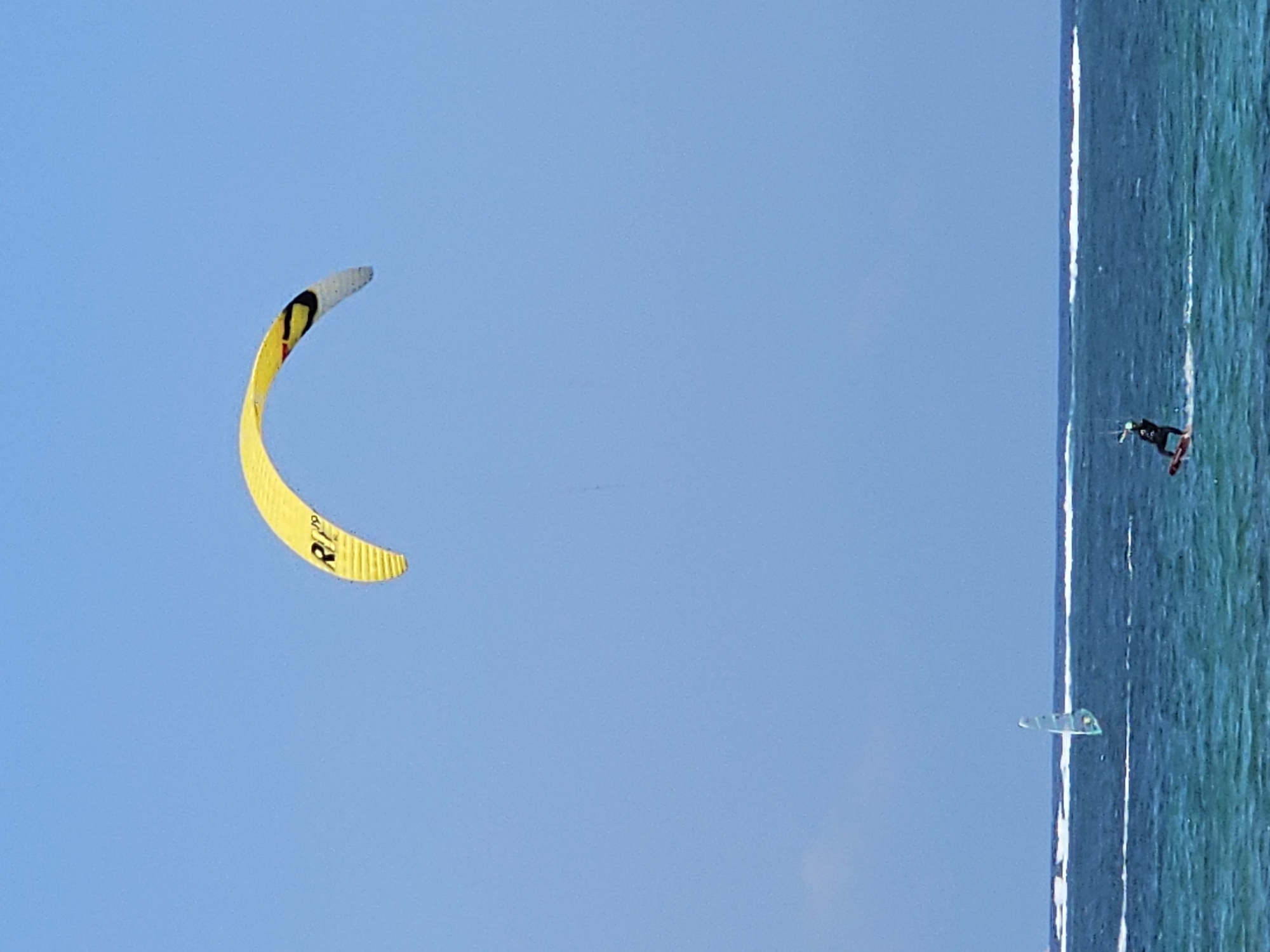 A kite surfer is on the water, performing a water sport. The kite surfer is wearing a black suit and a helmet, and is standing on a surfboard. The kite is yellow and white, with the logo "RRD" visible. The kite surfer is in the middle of the ocean, with a clear blue sky above. In the background, there is a windsurfer and a distant reef break. The water is choppy and appears to be a tropical location. The image was taken in the city of Denpasar, Indonesia.