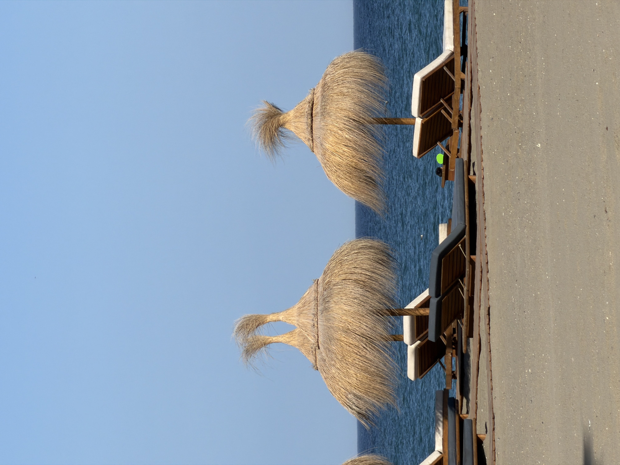 The image depicts a serene beach scene at Playa Del Sol-Villacana, Spain. In the foreground, a sandy beach stretches out, with several lounge chairs set up on wooden platforms. Two prominent straw umbrellas, with their distinctive conical shape and wispy straw details, provide shade over some of the chairs. The ocean, a deep blue, forms the background, meeting the clear blue sky at the horizon. The overall atmosphere is one of tranquility and relaxation, with the gentle sway of the umbrella straw suggesting a light breeze. There are no people visible in the scene.