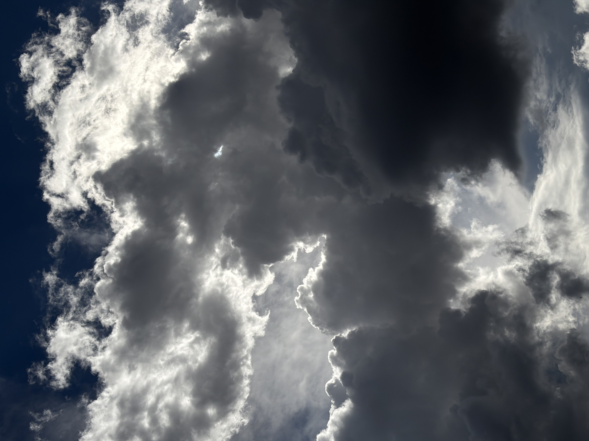 The image shows a sky filled with dark, heavy storm clouds. The clouds are predominantly gray and white, with some areas appearing nearly black. Sunlight breaks through in places, creating a sharp contrast between light and shadow and outlining the edges of some clouds with a bright glow. The sky itself is only visible in small patches, appearing as a deep blue. There is no indication of a specific location or cityscape.