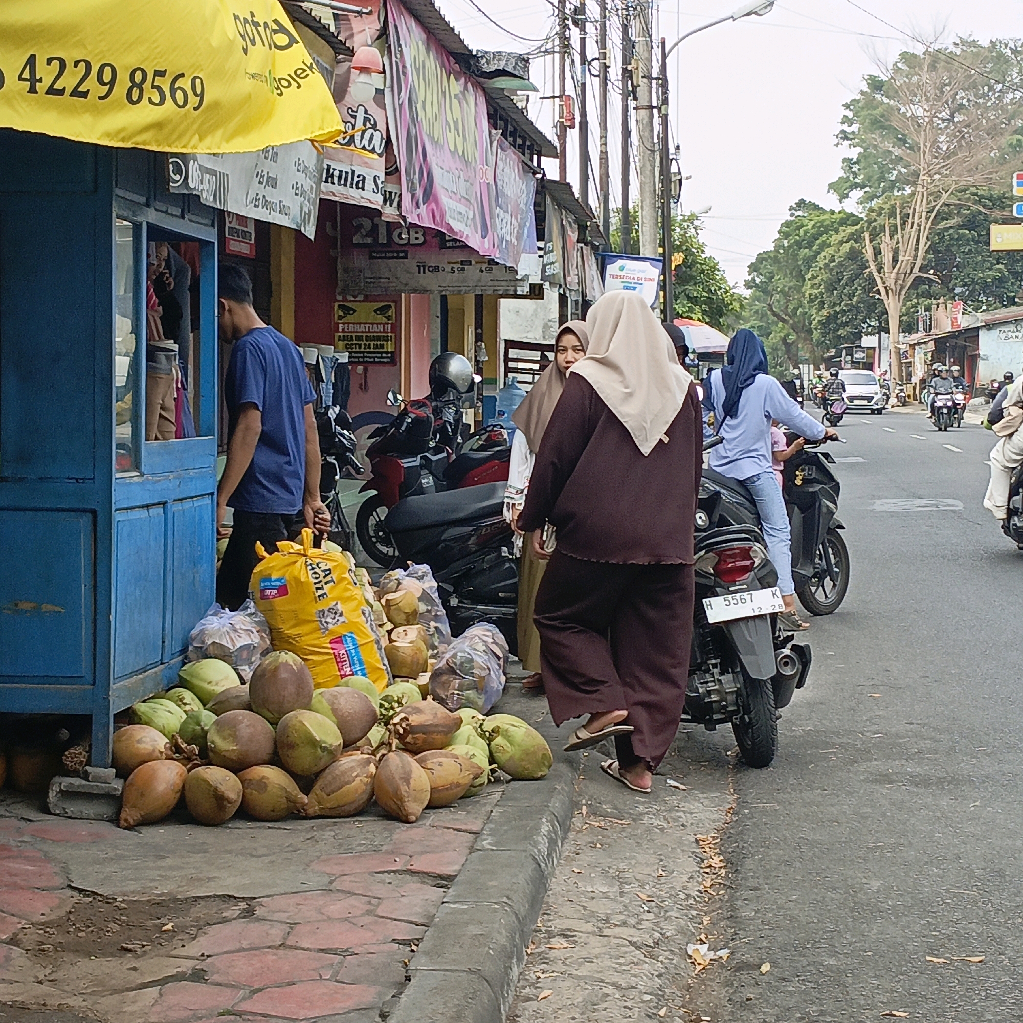 This is an outdoor scene in Salatiga, Indonesia, showing a street-level view of a roadside stall and adjacent street. In the foreground, a pile of coconuts is displayed on the sidewalk next to a blue wooden structure, possibly a kiosk or shop. A large yellow bag with "CAT HOZE" printed on it sits amongst the coconuts. A man in a blue t-shirt is seen from the back, walking away from the stall and carrying a plastic bag. To the right of the stall, three women are present; two are wearing hijabs, and one is partially visible behind the other. One woman is looking towards the camera, while another is seated on a scooter in the street. The street itself is paved and shows signs of traffic, with several motorbikes and a car visible in the background. Overhead power lines and streetlights are present, typical of urban Indonesian settings. The buildings lining the street have various signs and banners displayed, indicating commercial activity. The overall atmosphere suggests a typical day in a bustling commercial area of the city.
