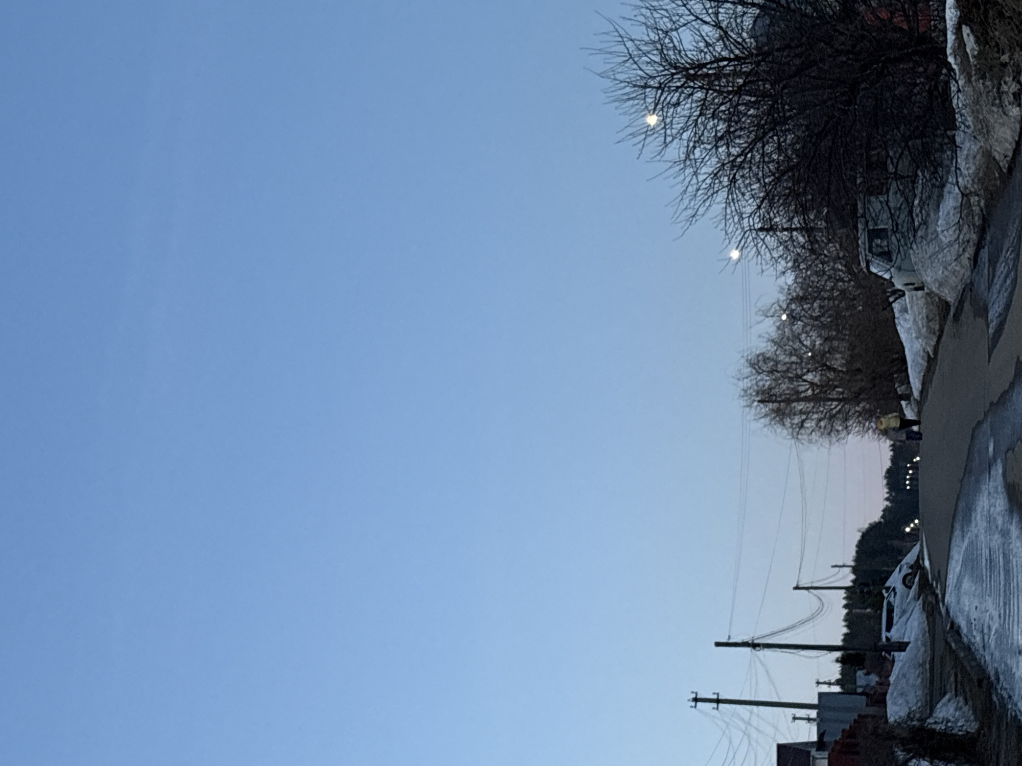 The image depicts a winter street scene in Прикамский, Russia. The sky is a pale blue. A row of bare trees lines the right side of the street. On the same side, a sidewalk is covered with melting snow and ice, next to a road where cars are parked. Utility poles with wires run alongside the street. Buildings are visible in the lower right corner, and in the distance, a dark strip of woodland appears against a lighter sky. There are lights on the poles near the end of the road.