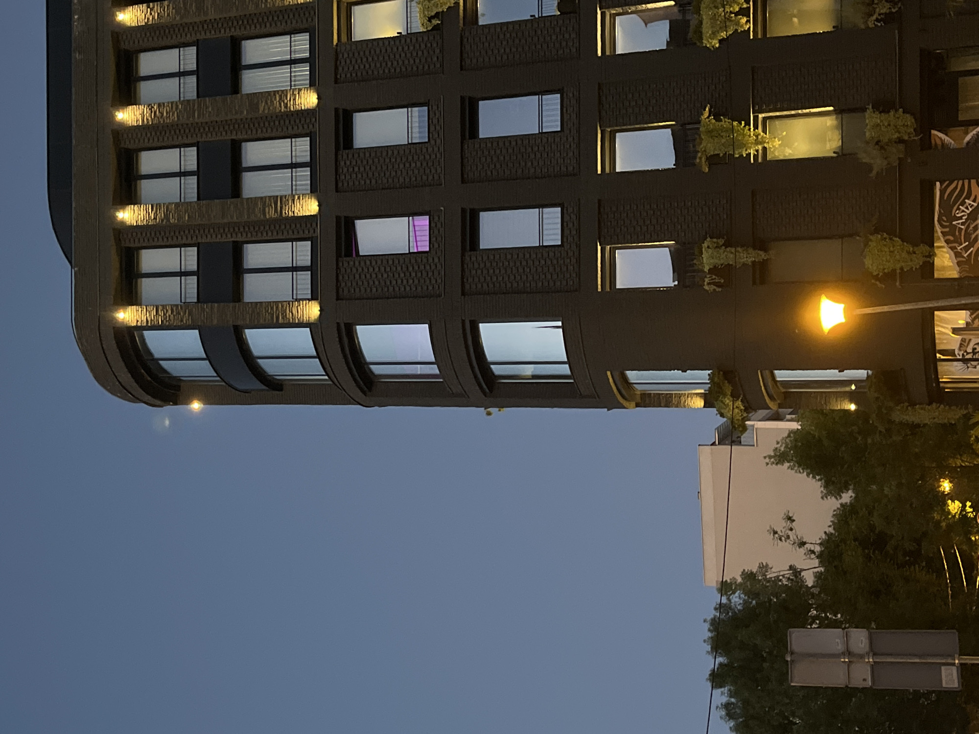 The image shows a multi-story building with a curved corner, situated in Belgrade, Serbia. The building has a dark, textured facade and evenly spaced windows, some of which are illuminated from within. There are small plants adorning some windows. A street light is visible, casting a warm glow. To the right of the building, part of a lighter-colored structure and trees are visible. At the bottom of the frame, there are road signs. The sky is a gradient of dark blue, suggesting it is evening or dusk.