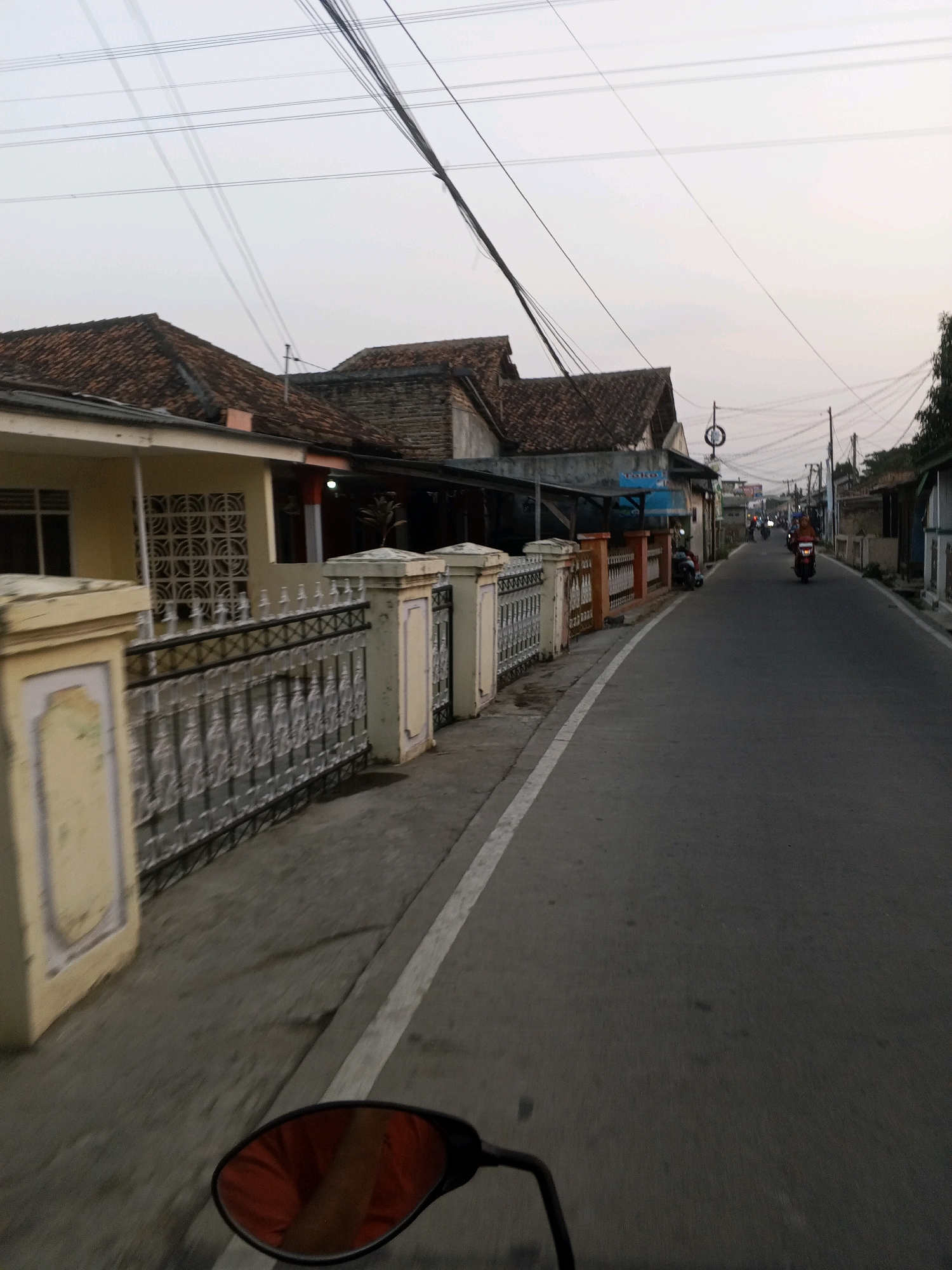 The image captures a street scene in Ciruas, Indonesia, likely during the late afternoon or early evening, given the diffused light and overcast sky. The perspective is from the viewpoint of someone riding a motorcycle, indicated by the motorcycle's rearview mirror in the lower left corner, reflecting a person wearing an orange garment. The street itself is paved and appears to be moderately trafficked, with several motorcycles visible in the distance.
Along the left side of the road, there is a low, decorative fence made of white-painted concrete pillars and black metal railing. Behind the fence, there are houses with tiled roofs and light-colored walls. Some houses have open verandas or covered walkways. Vent blocks are visible on one of the houses. Further down the street, more houses and small businesses line the road.
On the right side of the street, the road curves slightly to the right. More buildings, possibly residences or small shops, are visible. A few motorcycles are moving away from the camera, one with a rider wearing a reddish-orange headscarf and a light-colored shirt. Overhead, a complex network of power lines crisscrosses the sky, supported by poles. The overall impression is of a typical urban or semi-urban environment in Indonesia.