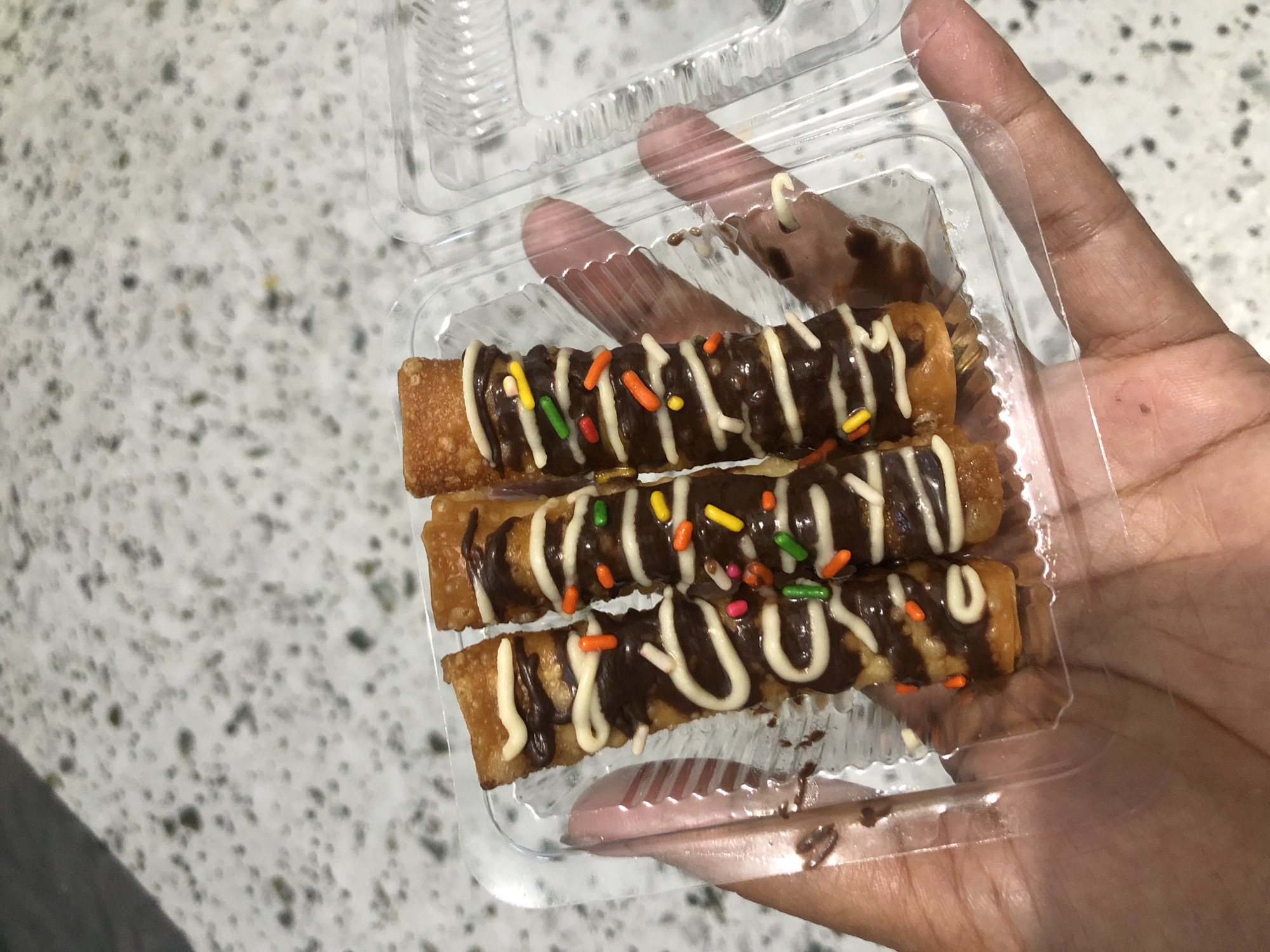 A hand holds a clear plastic container filled with three chocolate-dipped pastries, topped with white icing and colorful sprinkles. The pastries are cylindrical and appear to be fried, with a golden-brown exterior. The background shows a speckled countertop. The scene is indoors in Gampongasanlhoksukon Barat, Indonesia.