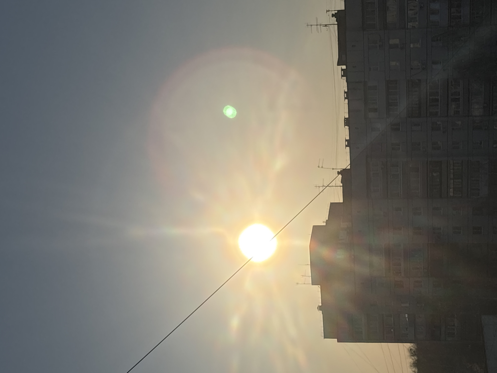 The image shows a daytime view of the city of Chaikovsky, Russia. The sun is low in the sky, casting a bright glare and lens flare. An apartment building is visible on the right side of the frame. A powerline cuts diagonally across the lower portion of the image. The sky is a hazy grey-blue.